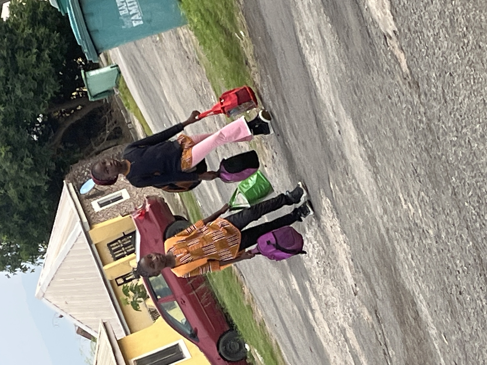 Two young people, likely children or teenagers, are walking on a paved road. The first individual is wearing a dark long-sleeved shirt and light pink pants. They are carrying a red bag with a long strap. The second individual is wearing an orange and brown patterned shirt and black pants. They are carrying a purple backpack. Both individuals appear to be wearing dark shoes.
Behind them, a red car is parked on the side of the road. Next to the car, a yellow house with a light-colored roof is visible. A large green trash bin with white text partially visible is also in the background, along with lush green trees and grass, suggesting an outdoor setting. The lighting suggests it is daytime, possibly morning or afternoon. The presence of bags and their attire might indicate they are going to or coming from school, or perhaps on an outing. The scene appears to be in a residential area. The text on the trash bin is partially obscured but includes the word "FAMILY".