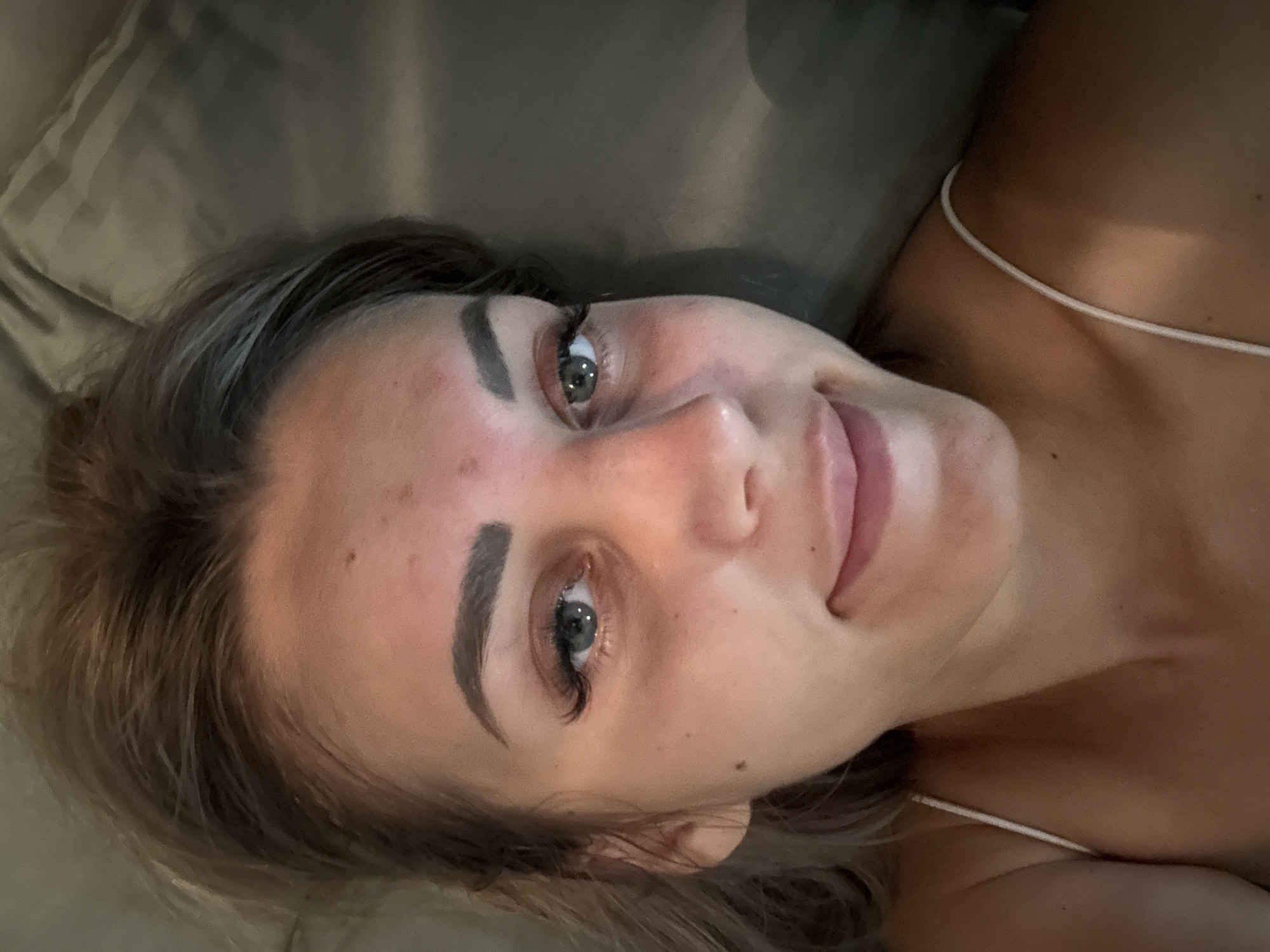 The image is a close-up selfie of a young woman with blonde hair and blue eyes. She is lying down, and her face is tilted towards the camera. She has on light makeup, including mascara and defined eyebrows. Her skin appears flushed, particularly on her cheeks and forehead. She is wearing a thin white strap top. The background is a textured, dark fabric, possibly bedding. The image does not contain any information that would allow for a determination of its location in Yuzhno-Sakhalinsk, Russia, or any other specific location.