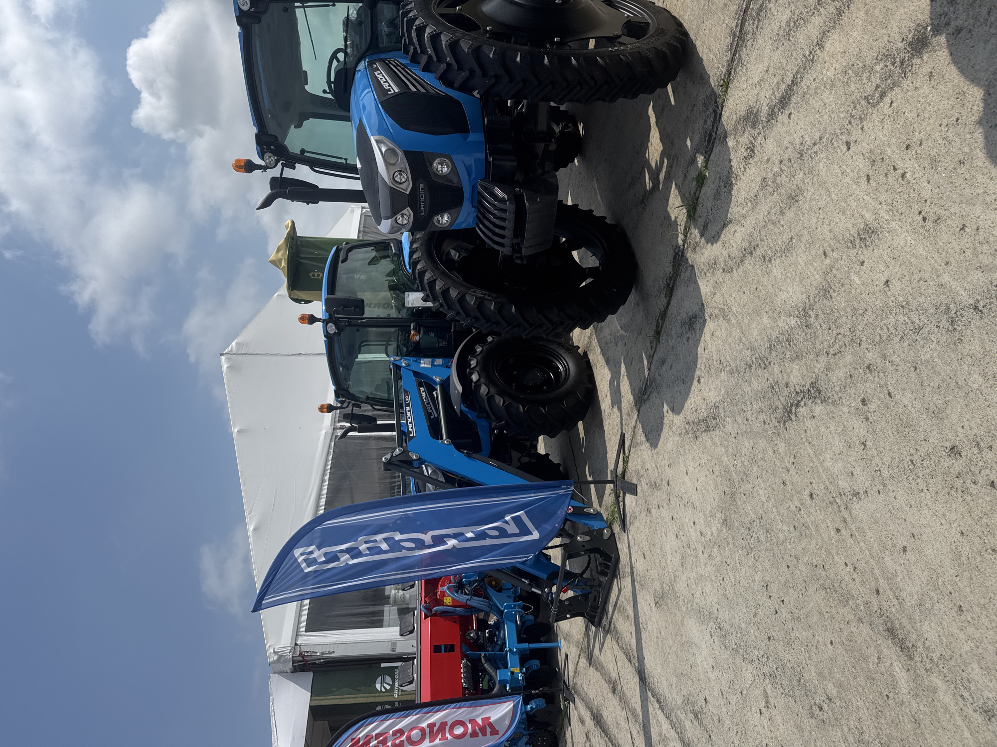 This image captures an outdoor display of agricultural machinery, predominantly blue Landini tractors, at what appears to be a trade show or exhibition. The scene is set on a concrete surface, with large white tents providing a backdrop.
In the foreground and midground, multiple blue tractors are visible. The largest tractor features the brand name "LANDINI" prominently on its hood and side, with "L20" also visible. Another smaller blue tractor, also branded "LANDINI," is partially visible behind it, equipped with a front loader. The tractors have large, rugged tires, typical for agricultural use.
A large blue flag, displaying the stylized "Landini" logo in white, is positioned in front of the smaller tractor and its attached implement, which appears to be agricultural equipment with blue and red components. Further in the background, a green piece of machinery with the text "KRONE" can be glimpsed, suggesting other brands are also present. A red and white banner, partially visible at the bottom left (and reversed due to the image orientation), shows the word "MONSOON."
The time of day is bright, sunny, and clear, indicated by the strong illumination and distinct shadows cast by the tractors and flag on the concrete. The sky is a clear blue with scattered white clouds, suggesting pleasant weather. The overall setting implies an event showcasing agricultural technology in an open-air environment, likely in Stara Zagora, Bulgaria, as per the provided context. No people are visible in the frame, allowing the focus to remain on the machinery and display setup.