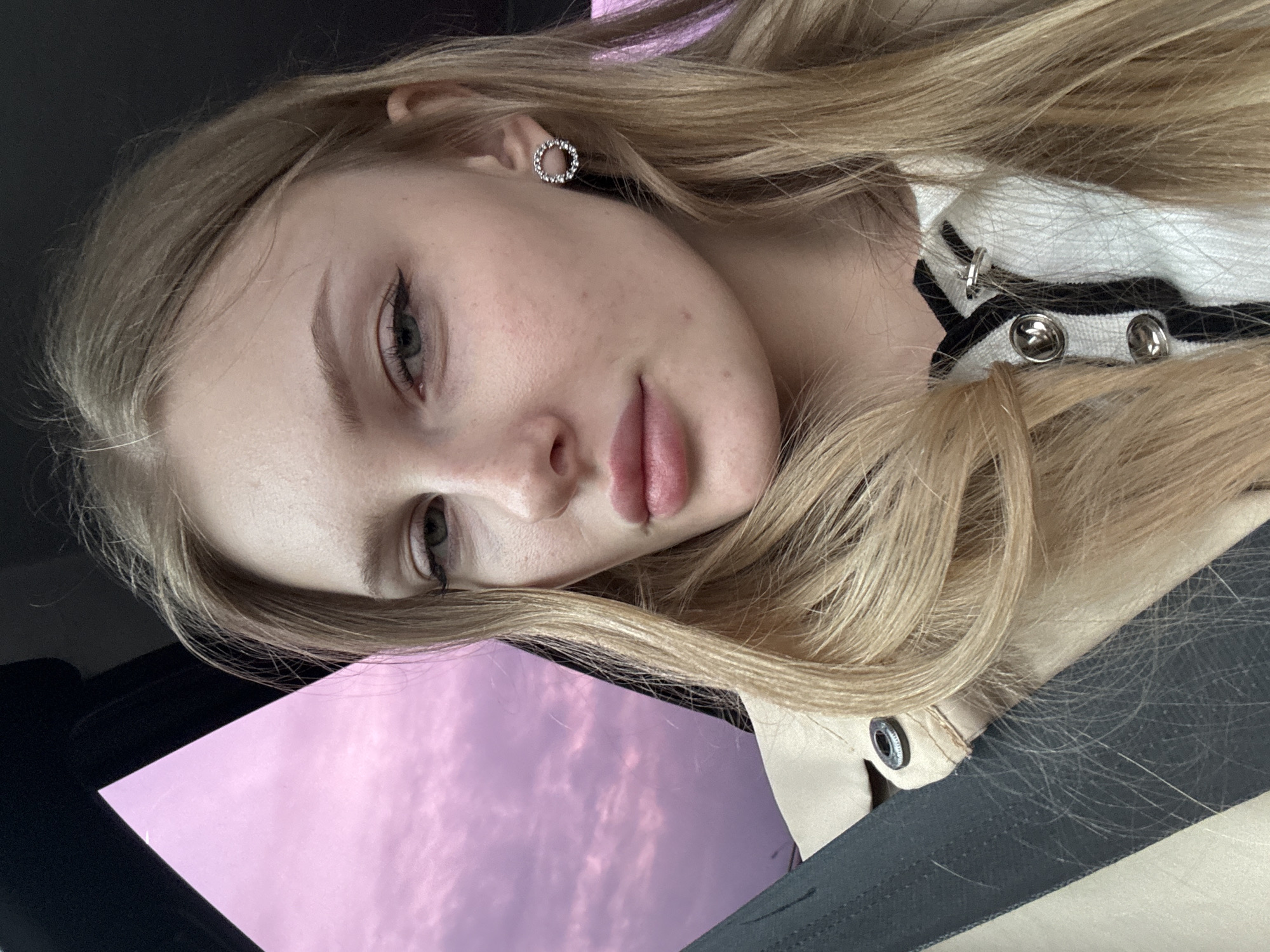 A young woman with light blonde hair and light skin is depicted in a selfie-style photograph. She has light eyes, full lips, and is wearing a circular earring. The backdrop visible through what appears to be a car window shows a pink-hued sky, possibly at dusk or dawn. The location is not discernible as being the city of Perm, Russia from the visual content of the image alone. The foreground includes parts of a jacket, a white shirt with black stripes, and the interior of the car. The woman's head is tilted, creating a diagonal composition.