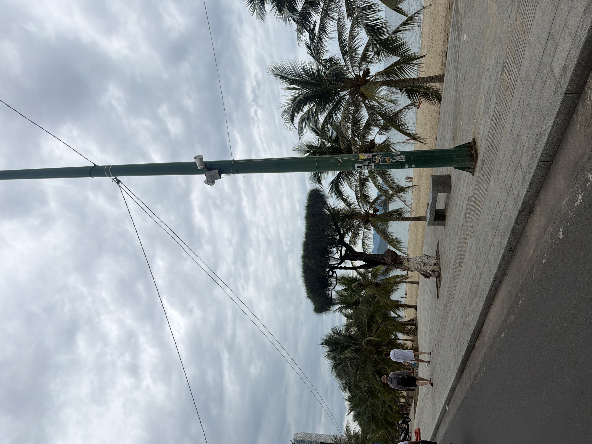 This image depicts a seaside promenade in Nha Trang, Vietnam, under a cloudy sky. The promenade is paved with grey stone tiles and runs parallel to a sandy beach. A green metal pole with two speakers mounted on it stands prominently on the promenade. Along the promenade, there are several palm trees, some with coconuts, and a unique tree with a rounded canopy. Two individuals are walking away from the camera on the promenade, one wearing a straw hat and a backpack, and the other in a white garment. Further down the beach, other people are visible. The sea is calm and extends to the horizon, where a hazy mountainous landscape can be seen. A modern building is visible in the background to the left, suggesting an urban environment.