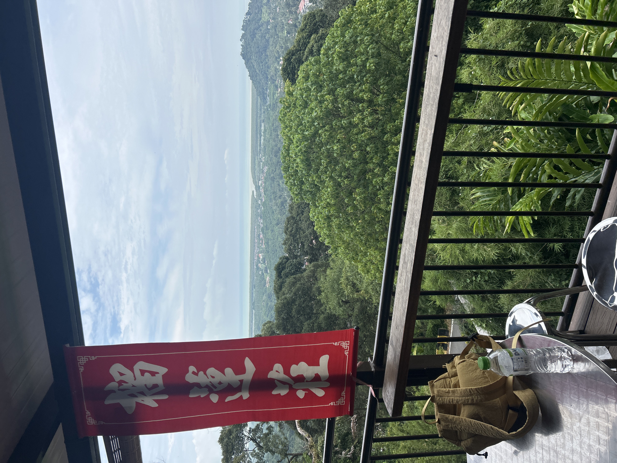 A red banner with white Chinese calligraphy hangs on the left side of a balcony overlooking a lush, green landscape. The banner features decorative border elements at the top and bottom. On a metal table on the balcony, a beige backpack and a bottle of water are visible. A metal railing runs along the edge of the balcony. The background reveals a dense forest, a body of water, and distant hills, suggesting a location in or near George Town, Penang, Malaysia, known for its hilly terrain and proximity to the sea. The sky is cloudy with patches of blue.
