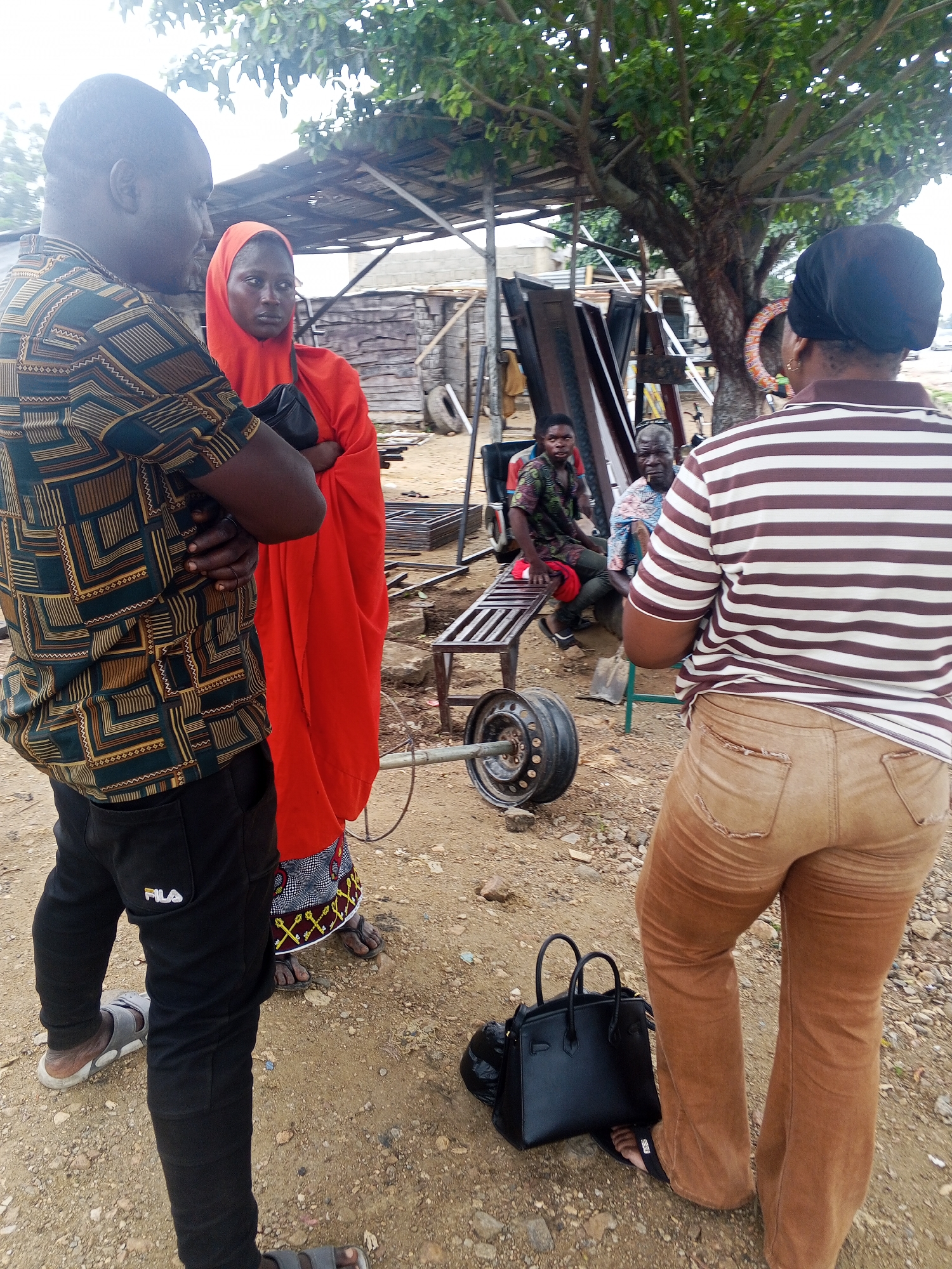 In Zango-ndaji, Nigeria, at an outdoor location with a dirt ground and some scattered rocks, a group of people are gathered near a makeshift structure with a corrugated metal roof and wooden supports. In the foreground, a man wearing a patterned shirt and black Fila pants stands with his arms crossed, facing away from the camera towards a woman in a vibrant orange headscarf and robe, who is also looking away. To the right, a person wearing a horizontally striped shirt and light brown corduroy pants faces away from the camera, with a black handbag at their feet. In the background, two men are seated on a metal bench. One man is wearing a patterned shirt and the other a blue and white patterned shirt, with a cigarette in his mouth. A barbell with a weight is lying on the ground in front of the bench, suggesting a possible outdoor gym setup or recreational area. The environment appears somewhat unkempt, with visible debris and unfinished construction elements in the background.