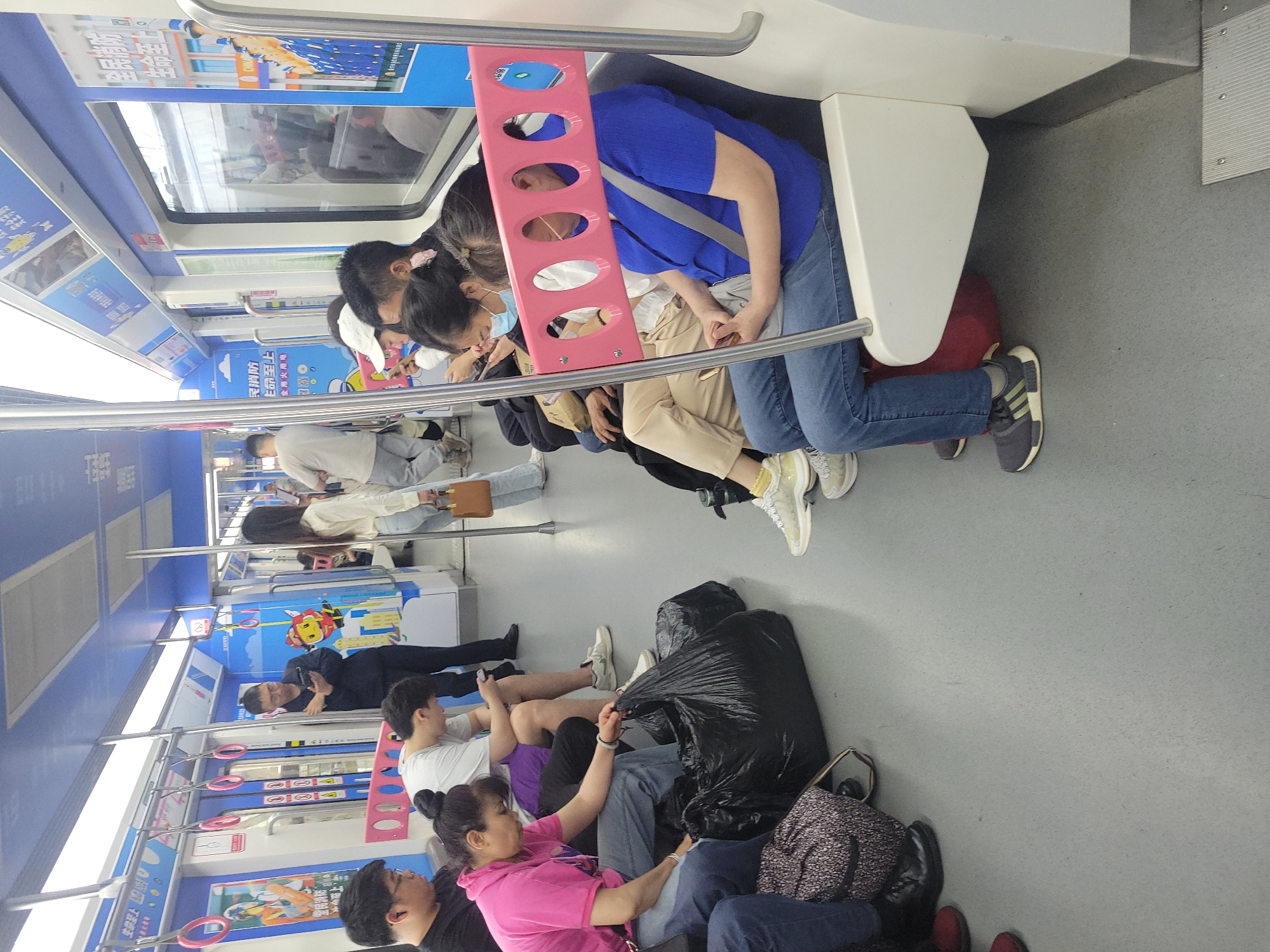 A train car in Liangjiang, China shows seated passengers. In the foreground, two people are seated on the floor, while others are seated on the benches. One person is wearing a pink shirt, and one person is wearing a white shirt. There is a large black bag next to the seated passengers. Advertisements on the walls include the phrase “生命至上 全民消防”.