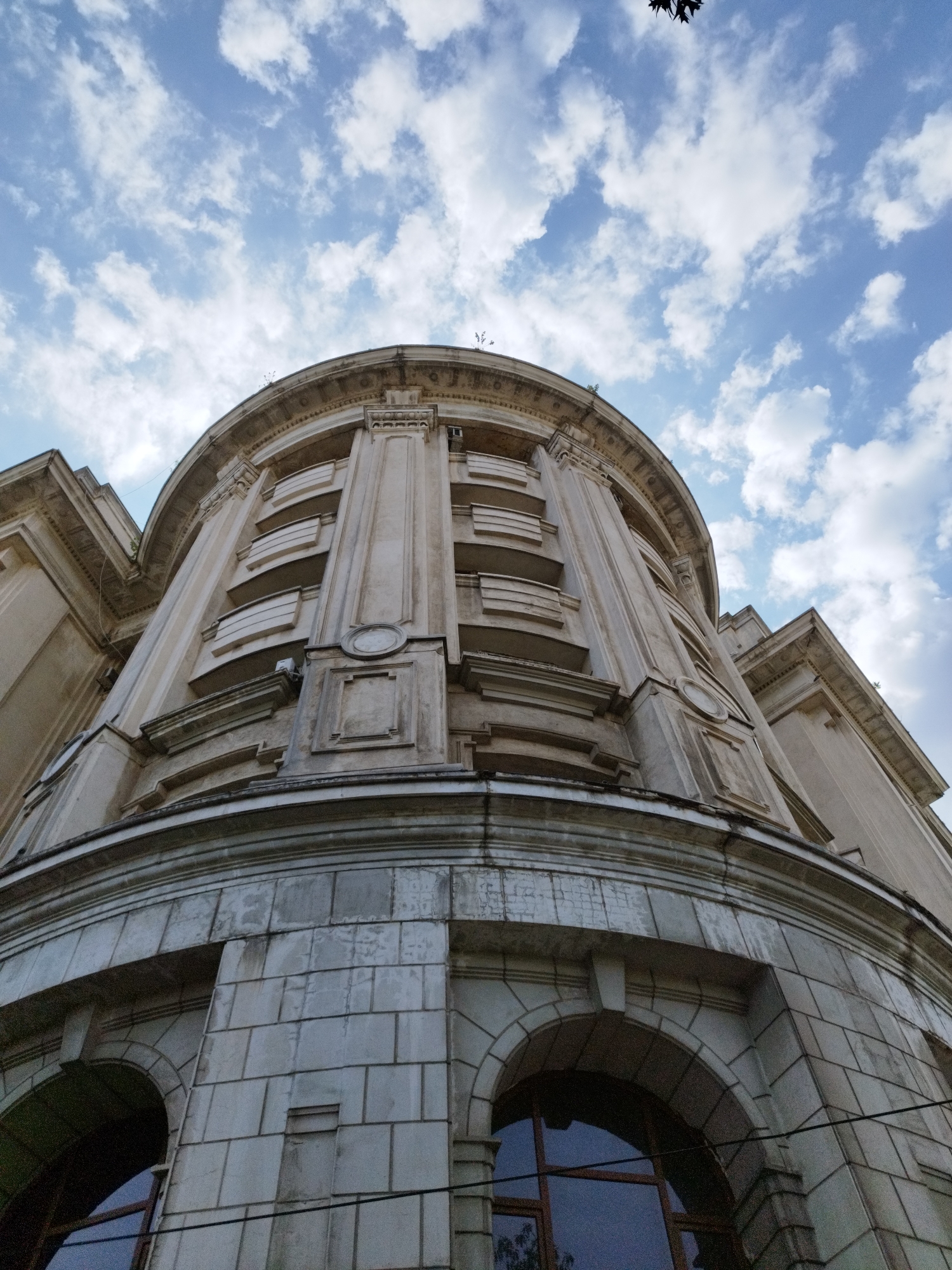 The image captures a low-angle view of the curved facade of a building, likely from Bucharest, Romania, against a backdrop of a bright blue sky with scattered white clouds. The building appears to be an older, grand structure, possibly with a historical or cultural significance. The architectural style features classical elements, including columns, pilasters, ornate moldings, and decorative recessed panels. The facade is constructed from stone blocks, with some areas showing signs of age and weathering. Large arched windows are visible at the lower level, reflecting the sky. The upper levels display a series of recessed balconies or window openings with simple horizontal detailing. A few dark tree branches are visible against the sky in the upper right corner, suggesting the presence of trees nearby. The overall impression is one of stately architecture set against a dynamic sky.