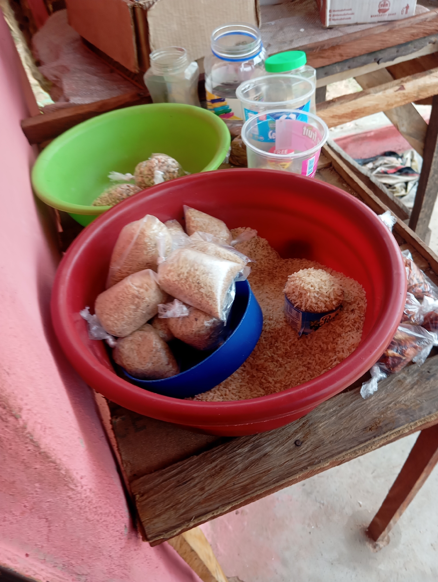 This image depicts a close-up, overhead view of a collection of rice stored in containers. In the foreground, a large red plastic basin is filled with uncooked rice. Several smaller, clear plastic bags, also containing rice, are scattered within and around the basin. A blue plastic scoop is partially submerged in the rice in the red basin. To the left of the red basin, a smaller green plastic basin holds a few more of the clear plastic bags of rice. In the background, various plastic bottles and containers are visible, some with lids. The scene appears to be outdoors, possibly a market stall or storage area, given the rough wooden surfaces and the presence of what looks like a pink concrete wall on the far left. The lighting suggests daytime. This scene is located in the city of Jalingo, Nigeria.