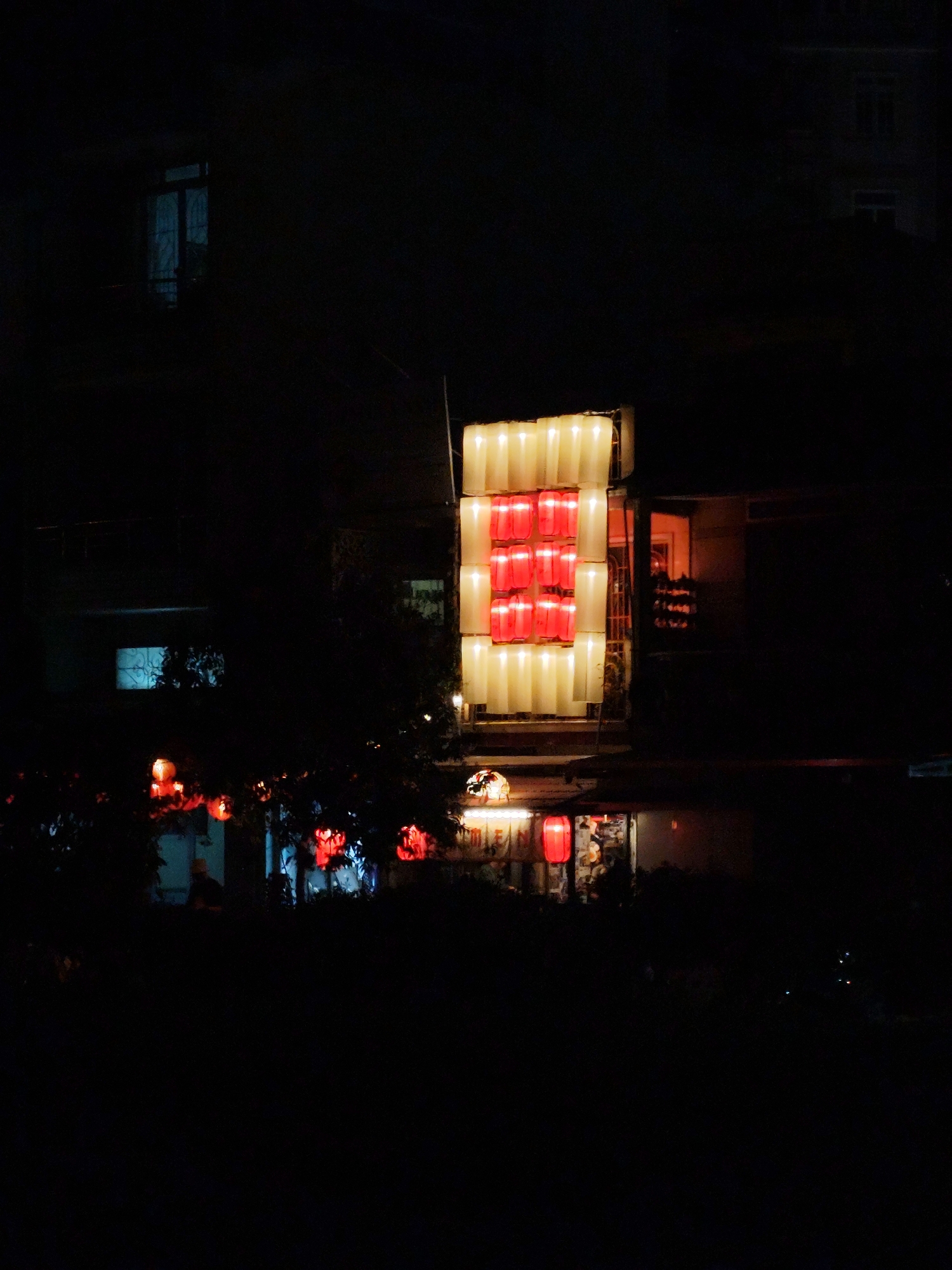 At night, the shot captures a scene in Da Lat, Vietnam. A structure of illuminated square lanterns stands out, some glowing white and others red, forming a distinct rectangle in the center of the frame. The lantern display is attached to a building. Further down, there are smaller red lanterns strung between trees, their soft light partially obscured by foliage. In the background are silhouettes of other buildings with a few visible windows. The overall scene is shrouded in darkness.