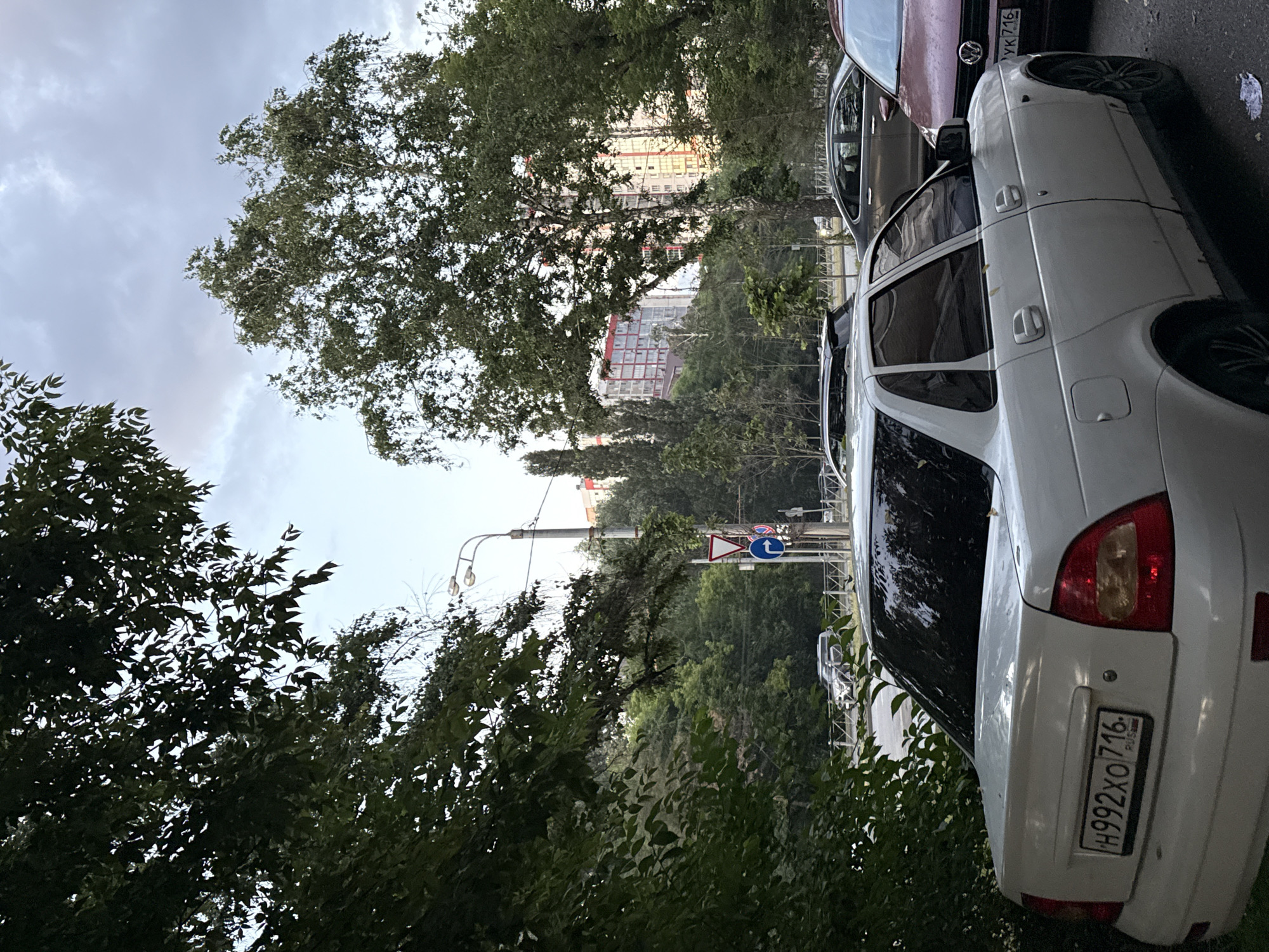In Kazan, Russia, on a street with trees lining the left side, a white sedan is parked. The car has a license plate that reads "H992XO 716". To the right of the white sedan, a maroon car is parked, with its license plate visible as "YK 716". In the background, there are trees and tall residential buildings with red and white facades. A street light and traffic signs are visible along the road. The sky is overcast with clouds.