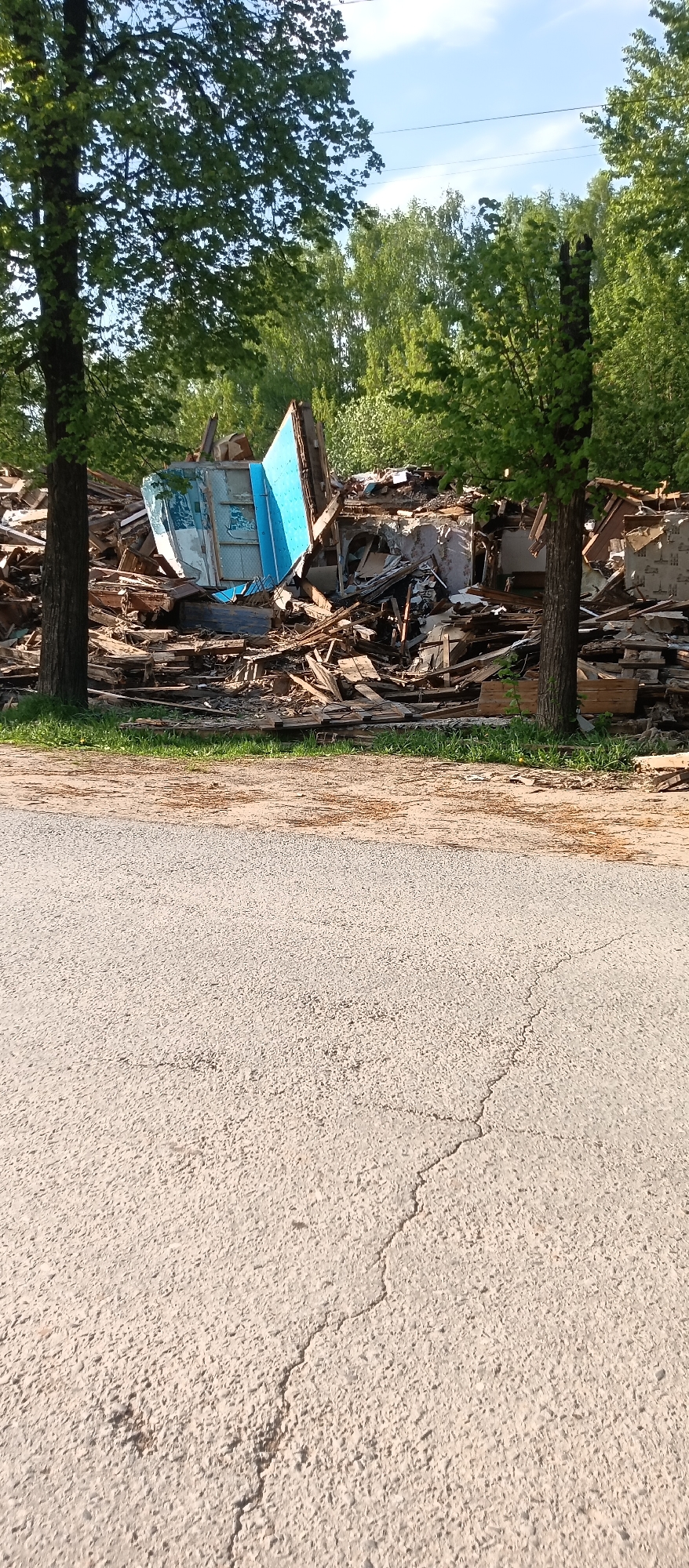 A debris pile is visible, possibly from a demolished building. The pile contains wood, structural elements, and fragments of what appears to be a blue wall or door. Two trees are positioned in front of the pile, partially obscuring the debris. In the foreground, a section of cracked asphalt pavement is visible, suggesting the pile is located near or adjacent to a road. The setting seems to be an urban or suburban area, given the presence of the road and surrounding vegetation. The location is Unknown, Unknown.