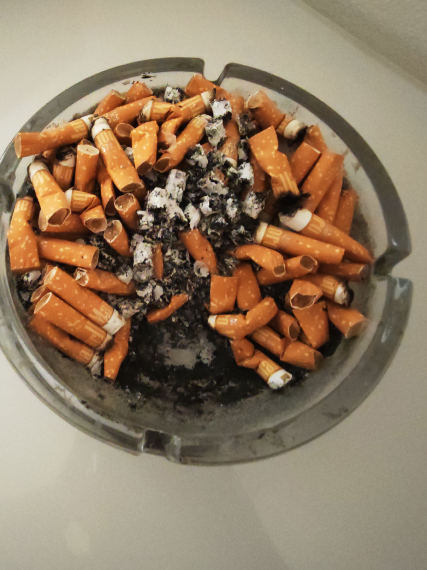 A close-up, high-angle shot shows a clear glass ashtray filled with numerous cigarette butts and ash. The ashtray is round with a ribbed edge and an indentation for resting cigarettes. The cigarette butts are orange and white, with many showing evidence of having been smoked, including black ash and frayed paper. The ash is gray and black, accumulating in the center of the ashtray. The surrounding environment is a plain, light-colored surface, possibly a table or countertop, and the background is a soft, neutral color. There are no people or specific landmarks visible that would indicate a location in Pilsen, Czechia. The scene is static, with no ongoing actions or interactions.