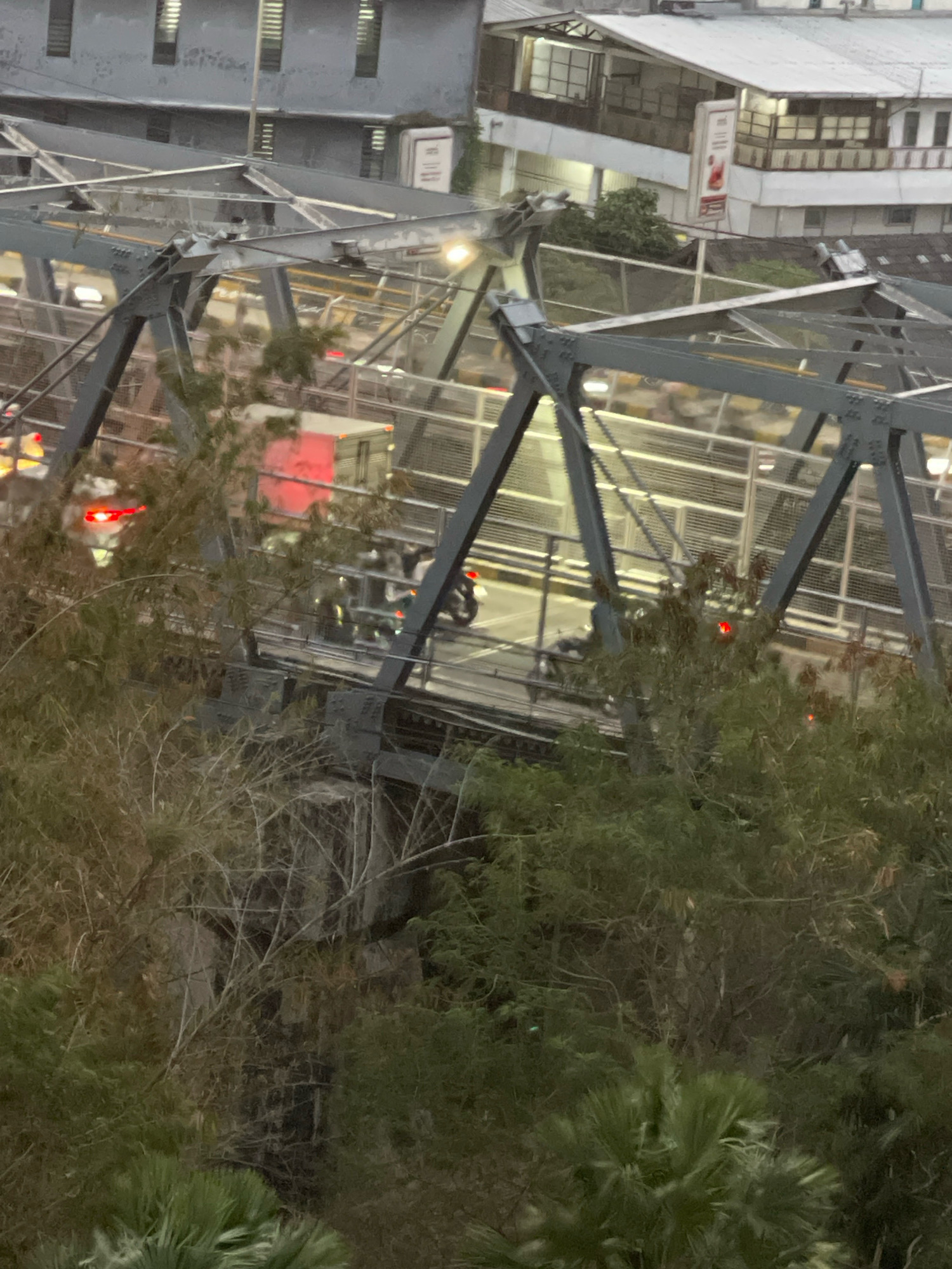 This is a medium shot, horizontal, eye-level, slightly blurry, outdoor, medium-light photograph taken from above looking down at a busy intersection on a bridge. The bridge itself is a truss bridge made of dark gray metal. The deck of the bridge is visible, with a yellow line separating lanes of traffic. There are multiple vehicles on the bridge, including at least two cars with their brake lights on, a white truck with a red taillight, and several motorcycles. Some of the motorcycles appear to have riders. There are also some pedestrians visible on the bridge.
The environment surrounding the bridge appears to be urban, with buildings visible in the background. The buildings are multi-story and appear to be made of concrete and metal. The sky is overcast and the lighting suggests it is either dawn or dusk. There are some trees and foliage in the foreground, obscuring some of the view. The overall impression is one of a bustling city scene with traffic in motion.