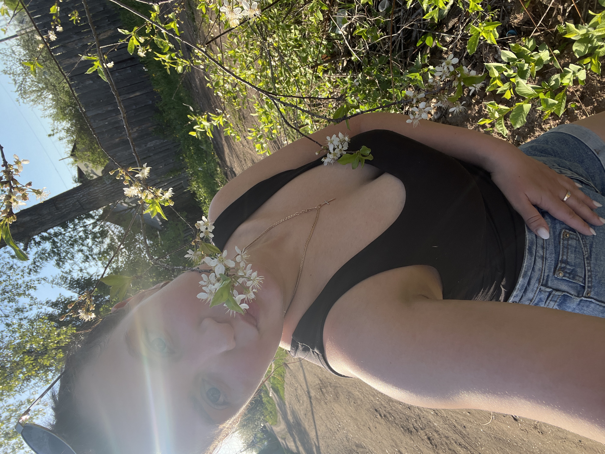 A medium shot depicts a woman lying on the ground outside, possibly in the Russian city of Noviy. She wears a dark tank top and denim shorts. White blossoms are positioned near her face and chest. A gold necklace is visible around her neck. The background includes a wooden structure and greenery. The lighting suggests a sunny day.