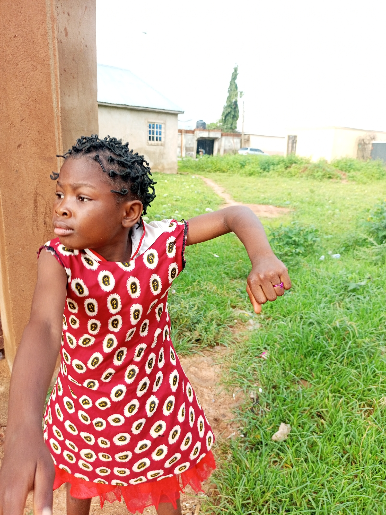A young girl with dark skin and braided hair stands outdoors, looking to her left with a slightly surprised or curious expression. She is wearing a red dress adorned with a white and yellow circular pattern. Her right arm is extended outwards, and on her right ring finger, she wears a thin, pink ring. The background consists of a grassy area with a dirt path winding through it. In the distance, there are buildings with light-colored walls and roofs, including one with a visible window. A tall, slender tree stands between two buildings, and a water tank is visible on the roof of a smaller structure. The sky is overcast and bright. The scene appears to be an outdoor location in a residential area, possibly in the city of Tempure, Nigeria, given the architectural style and vegetation.