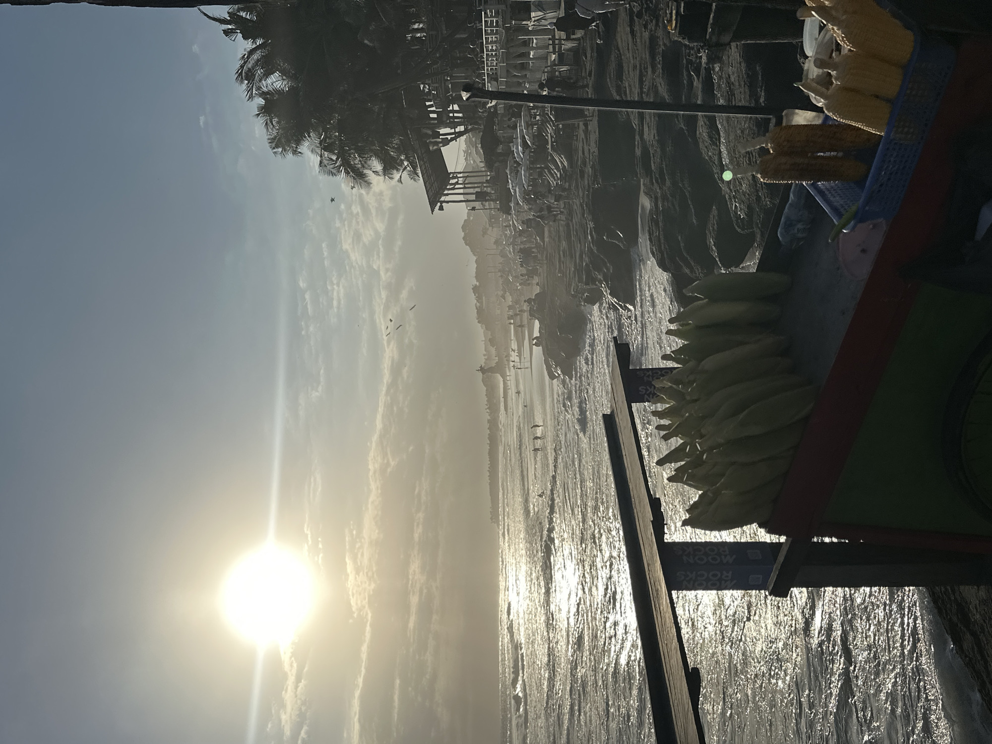 This is a photograph taken at the beach in North Kuta, Indonesia, during sunset. The sun is setting on the left side of the image, casting a warm glow and creating shimmering reflections on the ocean waves. The beach is populated with several people, some wading in the water and others walking along the shore. In the foreground, there is a food cart selling corn on the cob. The cart is colorful, with green and red sections. Several ears of corn are displayed on the cart, some raw and some roasted. Behind the food cart, there are rocky outcrops and a series of structures that appear to be beachside establishments, possibly restaurants or bars, with palm trees in the background. The sky is partially cloudy, with some streaks of light from the setting sun. The overall atmosphere is one of a lively beach scene at dusk.