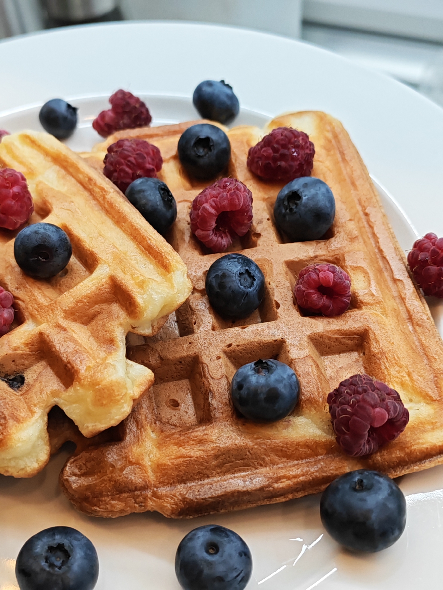 A close-up, overhead shot captures a plate of golden-brown waffles adorned with fresh blueberries and raspberries. The waffles are arranged on a white plate, with some overlapping. The berries are scattered across the waffle surfaces, with a few additional blueberries on the plate's surface. The lighting highlights the textures of the waffles and the vibrant colors of the berries. The background is blurred, suggesting an indoor setting, possibly a kitchen or dining area. The image does not contain any explicit location information for Bataysk, Russia.