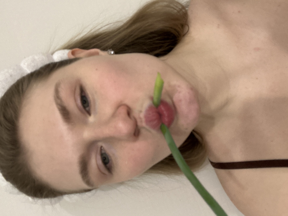 A woman lies on her back with her head turned towards the camera. She has light skin, light brown hair, and prominent eyebrows. She wears a white, frilly headband and a thin brown strap is visible near her shoulder. A green onion is positioned in her mouth, with the bulbous end appearing between her lips. The background is a smooth, light color. The image appears to be taken at a close-up angle, focusing on the woman's face and upper chest.