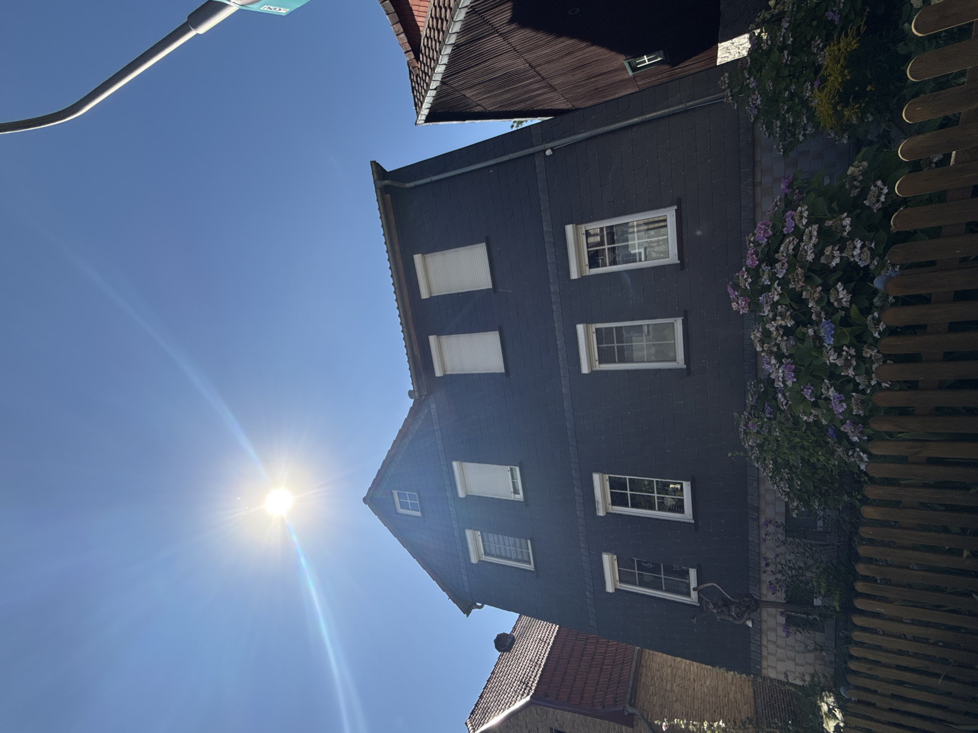 The image depicts the exterior of a multi-story residential building in Bad Sachsa, Germany, under a clear blue sky with bright sunlight causing lens flare. The building is clad in dark, slate-like shingles and features several white-framed windows. Some windows have white roller shutters partially or fully drawn. The roof is dark and steeply pitched. In the foreground, a wooden fence runs horizontally across the bottom of the frame, with flowering bushes, primarily hydrangeas in shades of purple and white, visible behind it. To the left of the main building, another building with a reddish-brown tiled roof is partially visible. To the right, a portion of a brown, vertically paneled structure is also in view. A street lamp is visible on the far right, extending upwards into the sky. The overall scene suggests a sunny day in a residential area.