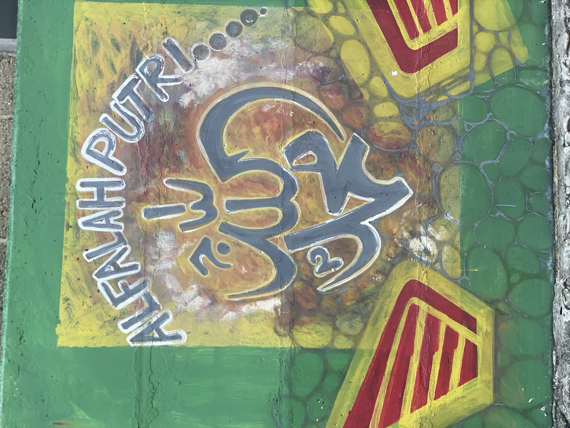 This is a photograph of graffiti art on a concrete wall in Banjarbaru, Indonesia. The artwork features Arabic calligraphy in grey and white, surrounded by yellow and orange paint. The calligraphy is in the center of the image and appears to be religious in nature. To the left and right of the calligraphy, there are red and yellow abstract shapes. The wall itself is painted green, with a textured pattern of darker green circles below the yellow and orange section. The top edge of the wall has a rough, grey concrete texture. The bottom of the image shows a cracked, grey concrete base. The overall scene is an outdoor urban environment.