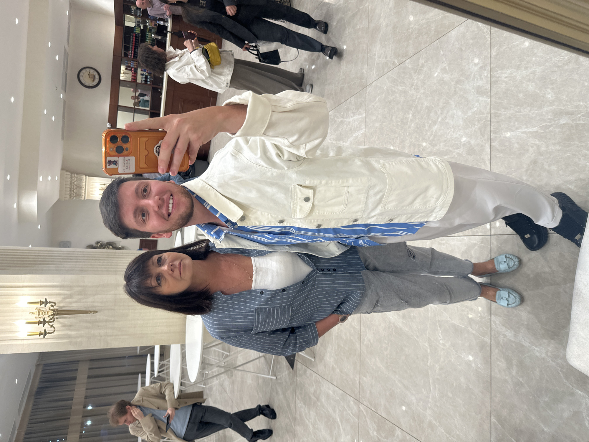 The image shows a couple taking a selfie in what appears to be an indoor establishment, possibly a restaurant or lounge in Moscow, Russia. The man in the foreground is holding a smartphone, which is angled to capture both himself and the woman beside him. He is wearing a light-colored jacket over a blue and white striped shirt. The woman is wearing a gray and white striped blouse over a white top, and gray pants. They are both smiling.
In the background, other individuals are visible, including a woman in a white jacket and a yellow bag, and a man in a darker outfit. There are also tables and chairs, suggesting a public space. The flooring is a light-colored marble or tile. A clock is visible on the wall in the background, indicating it is likely daytime. The overall mood seems relaxed and social. No visible text is present in the image.