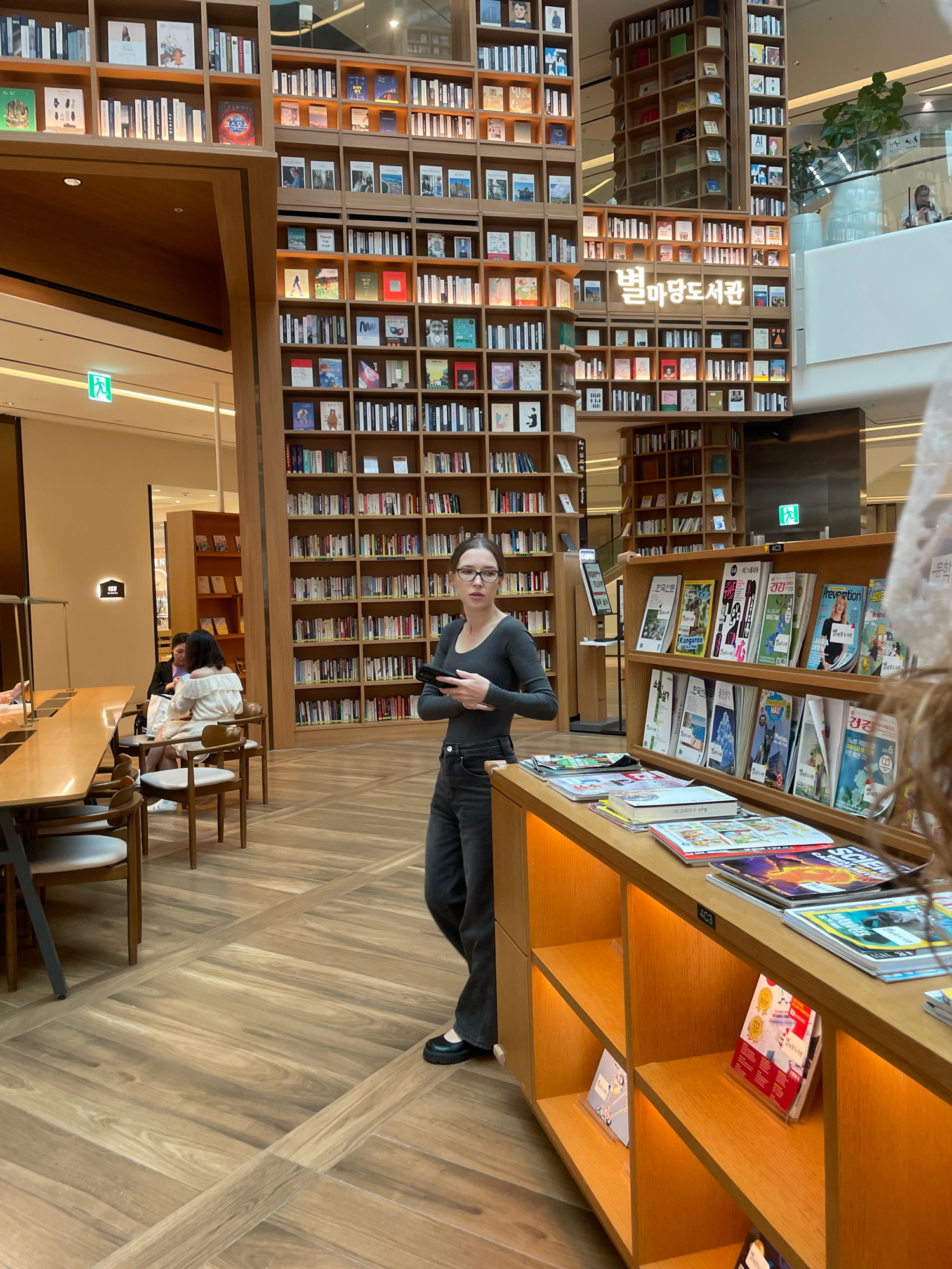 The image shows a woman with dark hair wearing glasses, a dark long-sleeved shirt, and dark pants standing in a modern library. She is holding a black phone in her right hand and is looking towards the camera. The library features tall wooden bookshelves filled with books and magazines. In the background, other people are seated at tables. A prominent neon sign with Korean text is visible on one of the bookshelves. The floor is made of polished wood planks arranged in a geometric pattern. The environment suggests a spacious and well-lit interior of a contemporary library. The location is identified as Starfield Library, located in Starfield Hanam, Gyeonggi Province, South Korea.