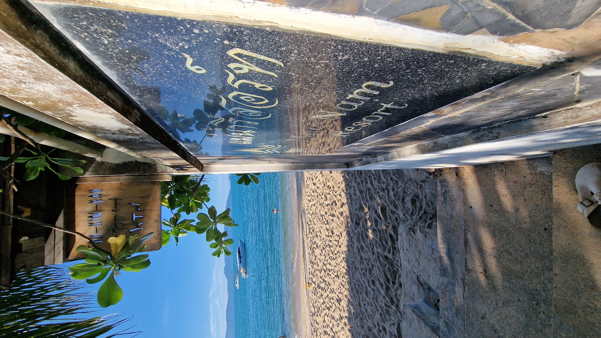 A resort sign stands on a stone wall overlooking a sandy beach and the blue ocean. The sign is made of black granite with Thai writing and the words "Nam Nam Resort" in English. A sandy beach leads to the ocean, where two boats are visible on the water. A person is swimming in the ocean. A pair of beige sandals are on the stone steps leading down to the beach. The sky is clear and blue. The resort appears to be located in Baan Mae Nam, Thailand.
