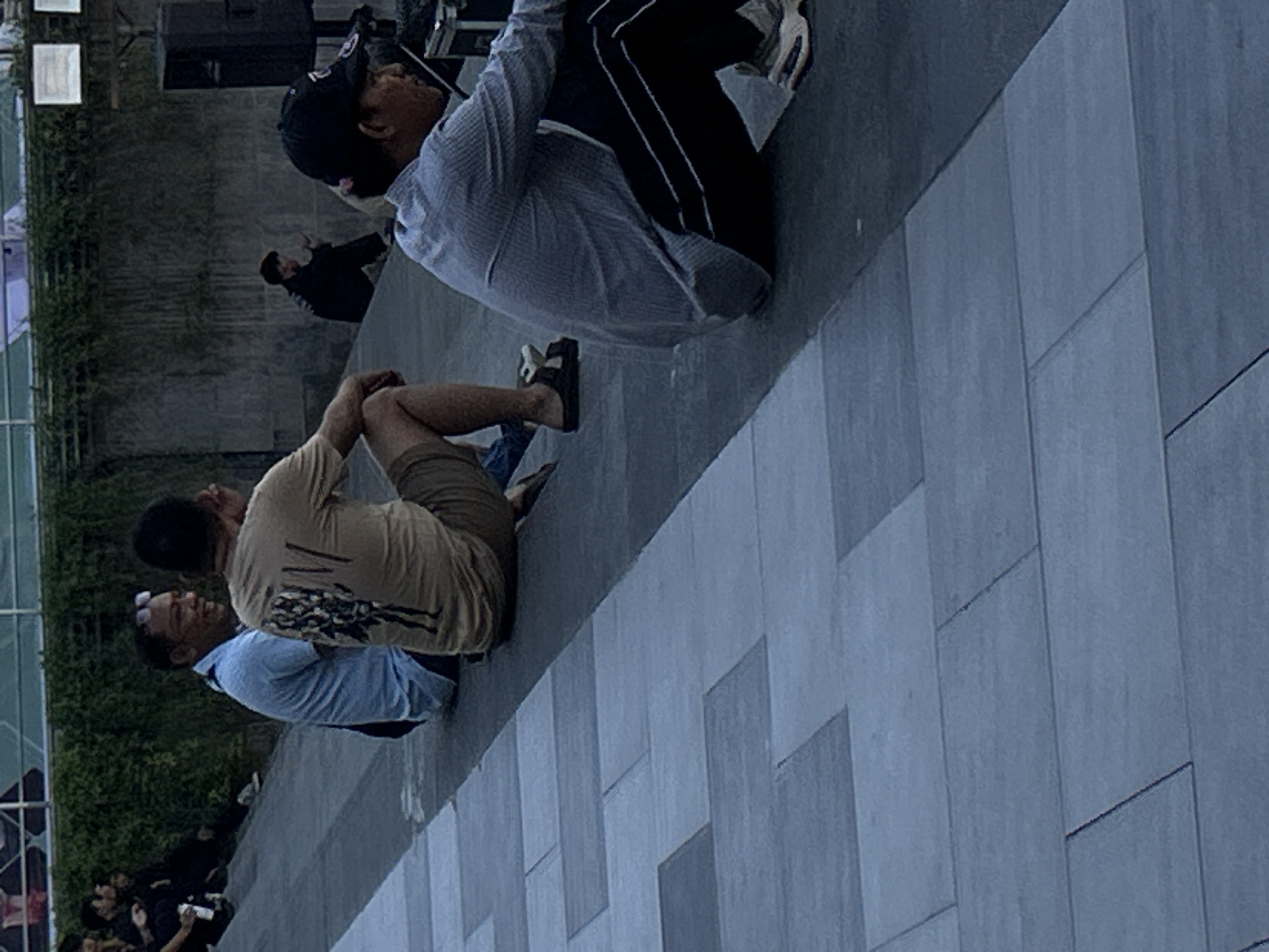 The image shows a group of people sitting on a tiered stone surface. In the foreground, a person wearing a blue and white striped shirt and black pants sits with their legs bent. To their left, another person in a tan t-shirt and shorts is also sitting. Further to the left, two men are seated on a higher tier; one is wearing a light blue shirt, and the other is out of focus. In the background, a person in a black jacket sits on a step. The environment appears to be an outdoor urban space with a stone wall and greenery. The scene is likely located in the city of Kuta, Indonesia, given the context of the prompt.