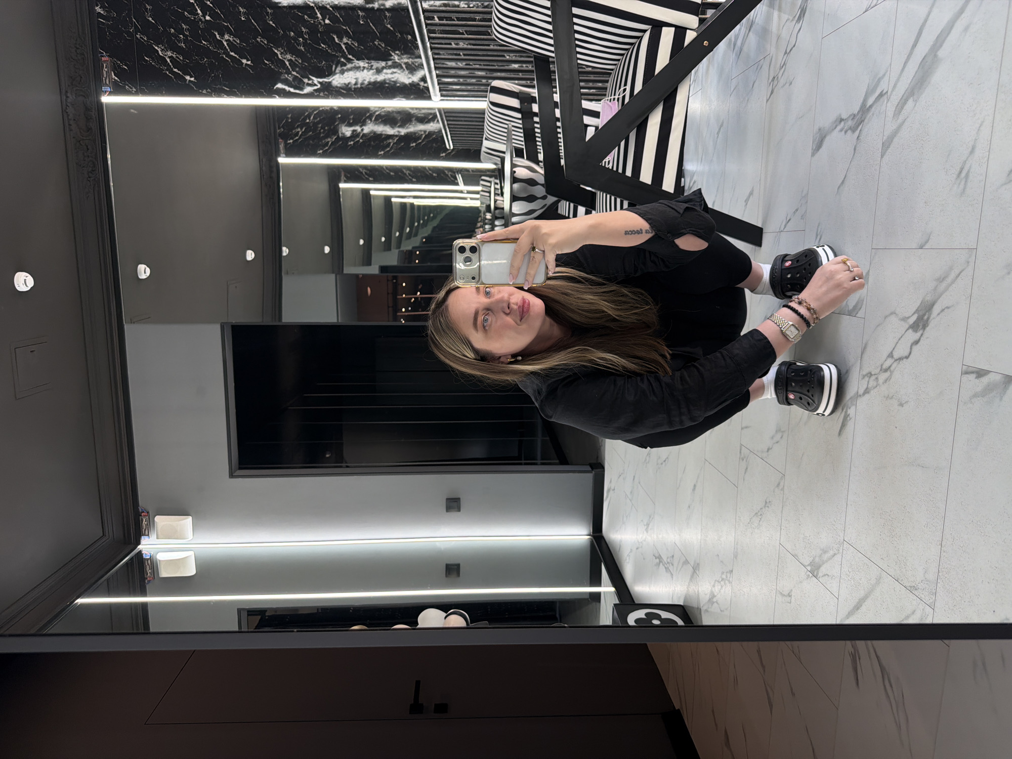 The image shows a woman in Moscow, Russia, taking a selfie in what appears to be a luxury boutique or salon. She is crouched down, holding a smartphone with a transparent case. She is wearing a black jacket, black pants, white socks, and black and white Crocs. She has bracelets and a watch on her left wrist. The background features a mirrored wall with linear lighting, creating an infinity effect. There is black marble wallpaper above the mirror. There is black and white striped furniture visible behind her. The floor is tiled with a light grey marble pattern. A product display shelf is visible along the bottom of the mirror, with some product packaging visible.