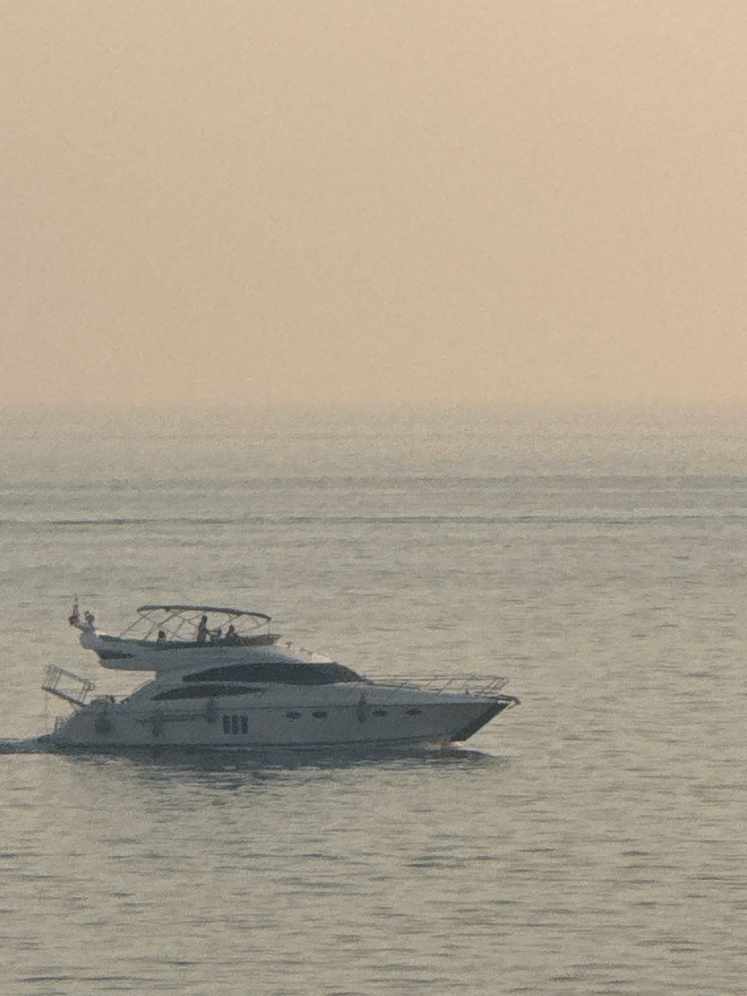 Two motor yachts are visible on the water. The larger yacht is in the foreground, moving from left to right, with its bow pointed towards the left. It is white with dark tinted windows and has a hardtop roof. Several people are visible on the deck of this yacht. A smaller yacht is visible behind and to the left of the larger yacht, also white and with a bimini top. The yachts are on a body of water that appears to be the sea, with a hazy horizon. The sky is a muted, peachy-orange color, suggesting either dawn or dusk. The location is the coast near Kamenovo, Montenegro.