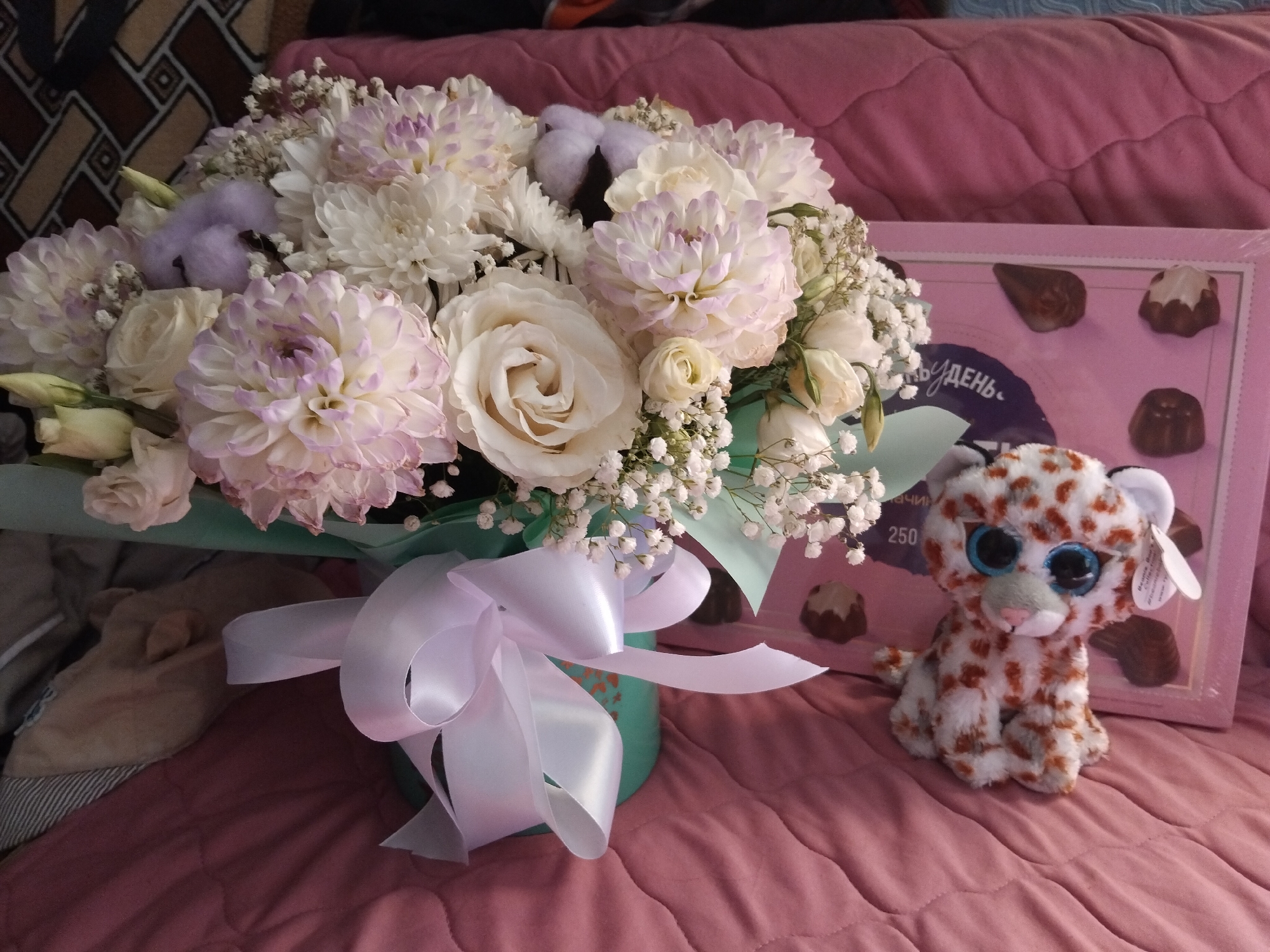 In Bila Krynytsia, Ukraine, a bouquet of white and pale purple dahlias and roses, accented with cotton balls and baby's breath, is presented in a teal cylindrical vase tied with a white satin ribbon. To the right of the bouquet is a pink box of chocolates with a clear lid, revealing several chocolate candies. A small, plush snow leopard toy with blue eyes sits in front of the chocolate box. The arrangement is placed on a pink quilted surface, likely a bed or sofa, with a patterned throw in the background.