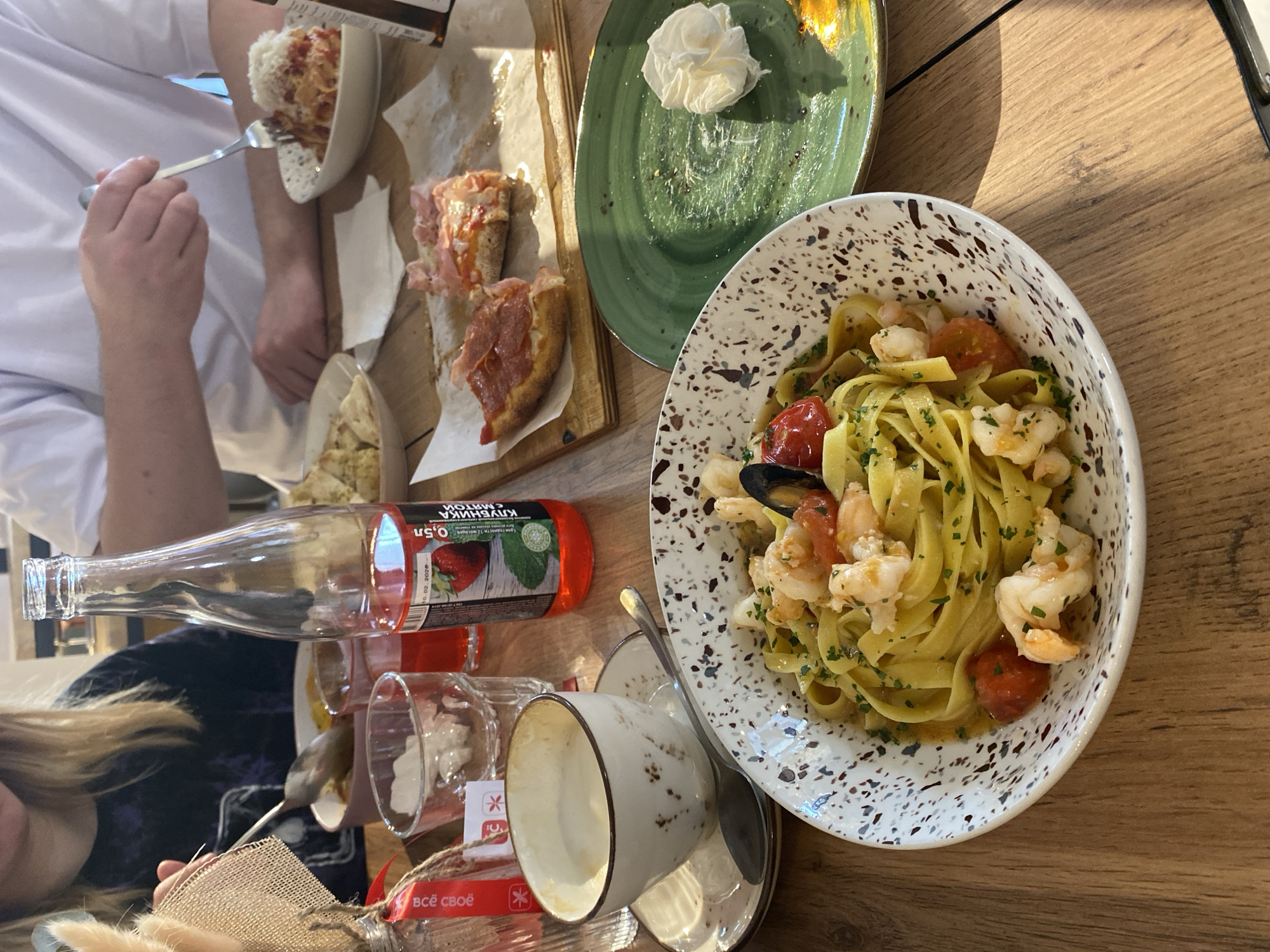 The image shows a table setting, presumably in a restaurant or cafe, with several food items. There is a bowl of pasta with shrimp and tomatoes, a wooden board with bread topped with cured meat, and a green plate with what appears to be whipped cream. A person is visible on the left side of the frame, eating from a bowl with a fork. There are also a bottle, glasses, a cup and saucer on the table. The location is Izhevsk, Russia, based on the inscription on the bottle. The scene depicts a casual meal being enjoyed at a food establishment.