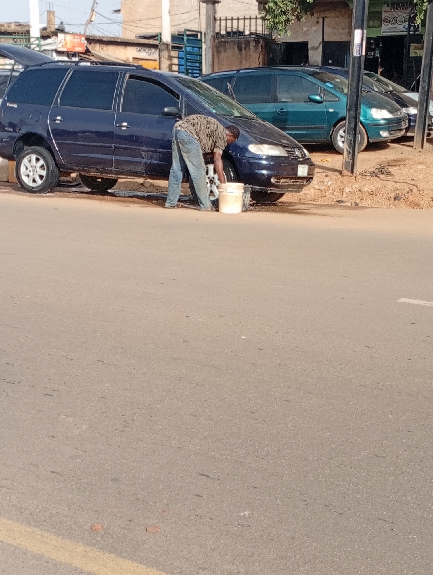 A person wearing a camouflage patterned shirt and blue jeans is washing the front right wheel of a dark blue minivan. The minivan has its rear hatch open. The person is using a white bucket and appears to be rinsing the wheel. Several other vehicles are parked nearby, including a teal colored minivan and a silver sedan. The scene is set on a dirt and asphalt roadside, with a paved road in the foreground. Buildings and overhead wires are visible in the background. The location is identified as Jos, Nigeria.