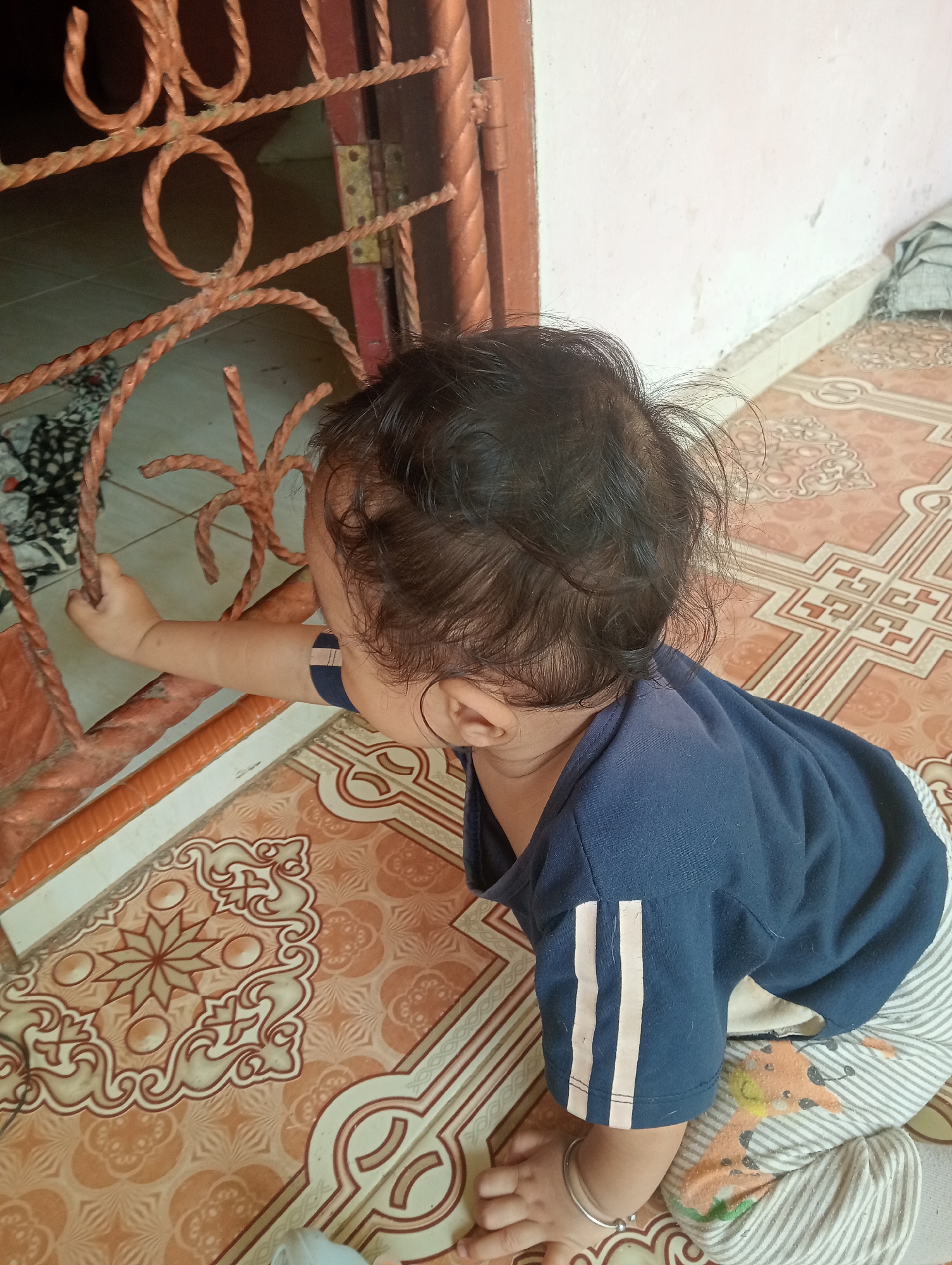 A young child, likely a toddler, is seen from a high angle, on all fours on a tiled floor. The child is wearing a dark blue shirt with white stripes on the sleeves and striped pants with a cartoon giraffe pattern. The child's right hand is reaching out towards a rusted metal gate with ornate circular patterns, appearing to hold onto it. The floor is covered with patterned tiles in shades of orange and cream, with a decorative border. In the background, a wall painted in a light color is visible, along with the edge of a closed door with metal hinges. The scene suggests an indoor or partially covered outdoor setting, possibly a home in Campago Barat, Indonesia. The child's focus is on the gate, implying curiosity or an attempt to explore. The lighting suggests it could be daytime.