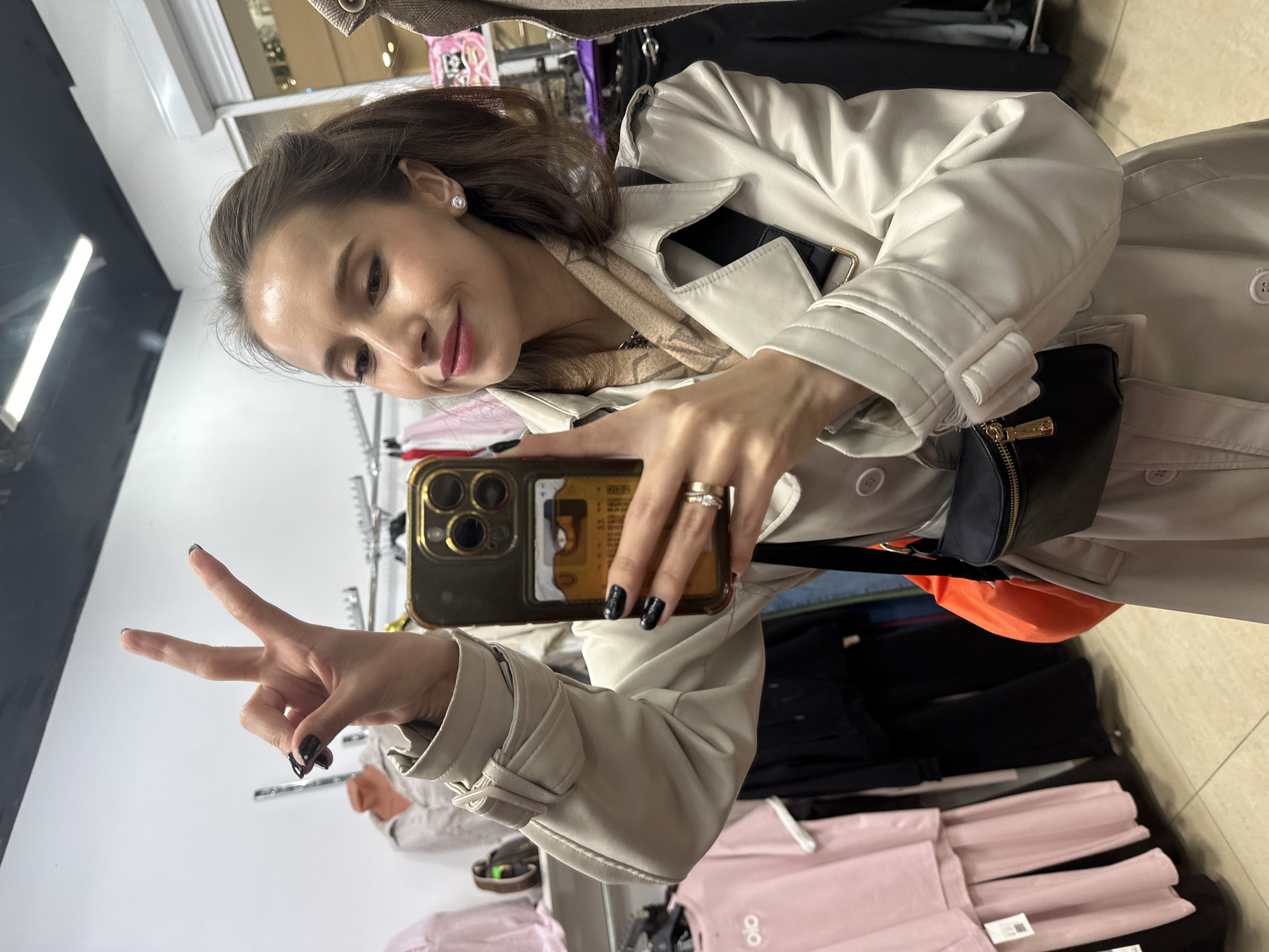 A young woman is taking a selfie in a clothing store. She is wearing a light-colored trench coat and holding her phone up, displaying a peace sign with her left hand. She is smiling at the camera. The background shows clothing racks with various items of clothing, mostly in pastel colors. A small black and gold purse is visible on her coat. The location appears to be indoors in a retail setting, likely a clothing store. It is not possible to determine the exact location in Yekaterinburg from the image.
