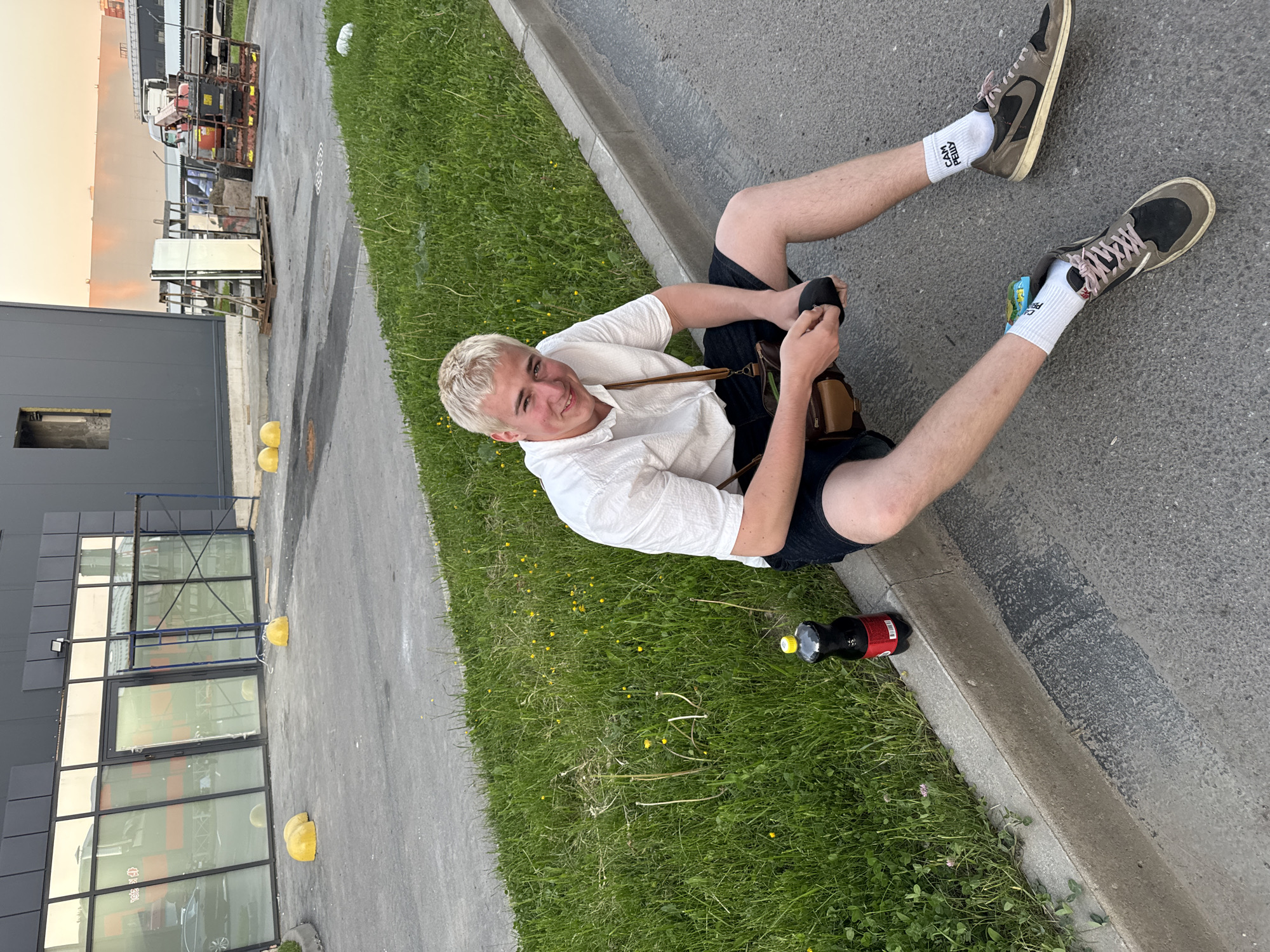The image depicts an outdoor urban or industrial scene during late afternoon or early evening in Unknown, Unknown. A young male with bleached blonde hair, wearing a white short-sleeved shirt, dark shorts, white socks, and dark sneakers, is seated on asphalt pavement adjacent to a concrete curb. He holds a black smartphone in his hands and is smiling directly at the camera. A brown leather messenger bag hangs from his shoulder. A dark-colored beverage bottle with a red label and yellow cap rests on the curb beside him. His right sock displays the Cyrillic text "САМ РЕШУ," and his left sock shows "ЭЙ ДВ." A small, colorful object is tucked into the laces of his left shoe.
To his left, a strip of green grass, containing small yellow wildflowers and some sparse areas, runs along the curb. In the background, a modern building with dark grey cladding and reflective glass windows is visible. A blue metal scaffolding structure stands outside the windows, and multiple yellow hemispherical concrete bollards are scattered on the pavement. Reflections in the windows show orange elements and a partial view of a dark vehicle. To the right, another dark-clad building, possibly under construction, features a rectangular opening. Further back, a wooden pallet holds several large sheets of reflective material, possibly glass or mirrors, next to an industrial lifting machine or small crane, colored red and rust-brown. A large, light-colored building is visible in the distant background under warm, low-angle sunlight. The asphalt pavement shows wear and some stains.