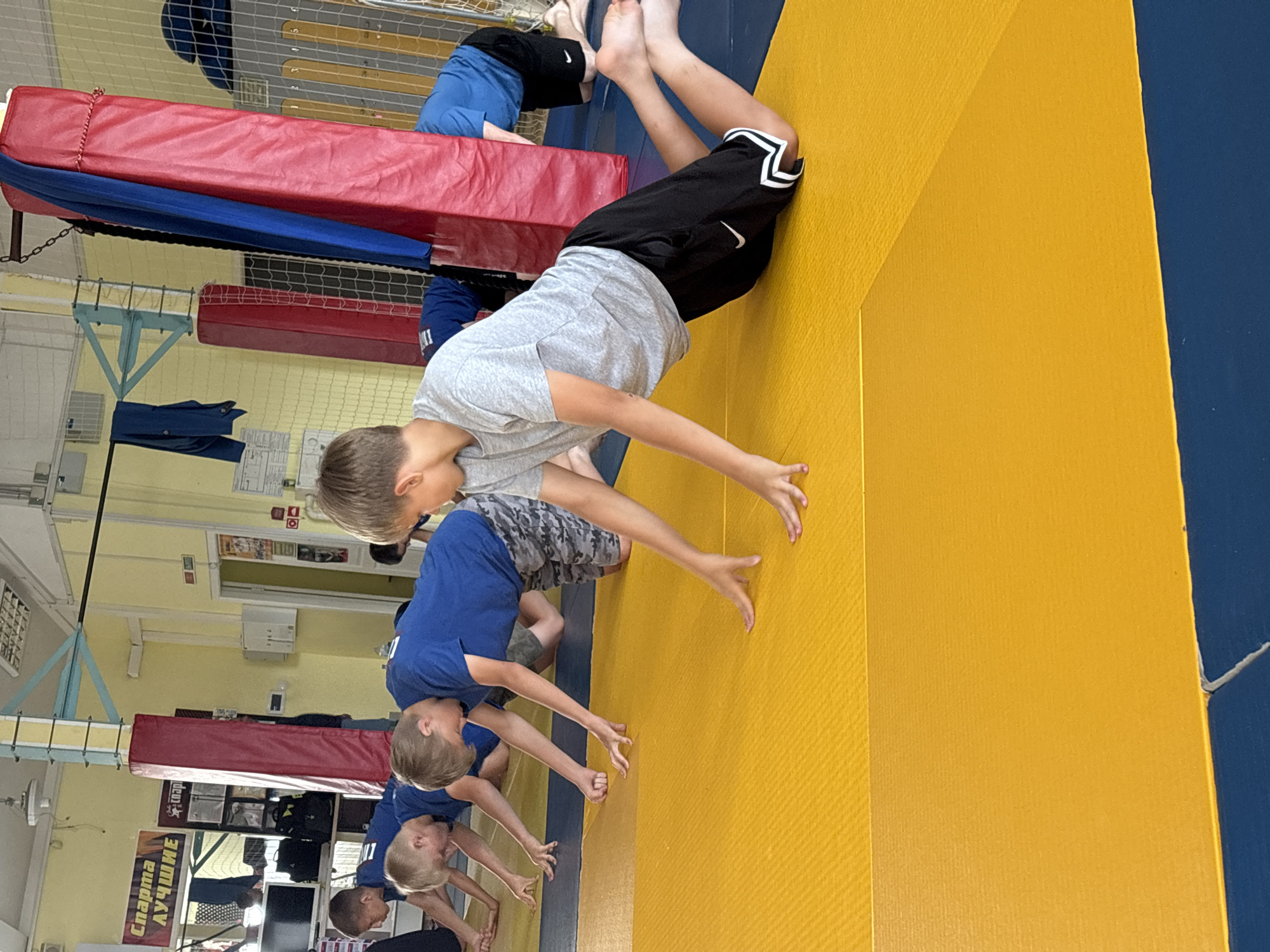 The image depicts a group of young boys engaged in physical training on a wrestling mat in an indoor gymnasium. The gymnasium appears to be located in Chaikovsky, Russia, as indicated by a banner in Russian that translates to "Sports for the Best." The boys are on a yellow and blue mat, with some in blue t-shirts and others in a gray t-shirt and black shorts. They are in a prone position, with their hands on the mat, suggesting a warm-up or a specific exercise. Red and blue padded pillars are visible in the background, along with gym equipment and netting. The overall atmosphere suggests a sports training session for young athletes.