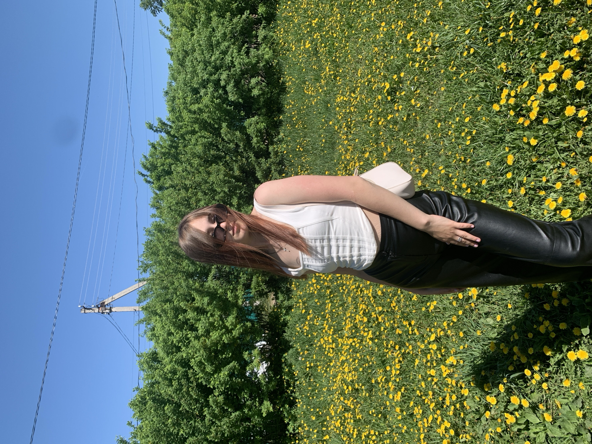 A woman is standing in a field of dandelions on a sunny day in Фоки, Russia. She is wearing black leather pants and a white top, and sunglasses. There are trees in the background and a power line pole to the left.