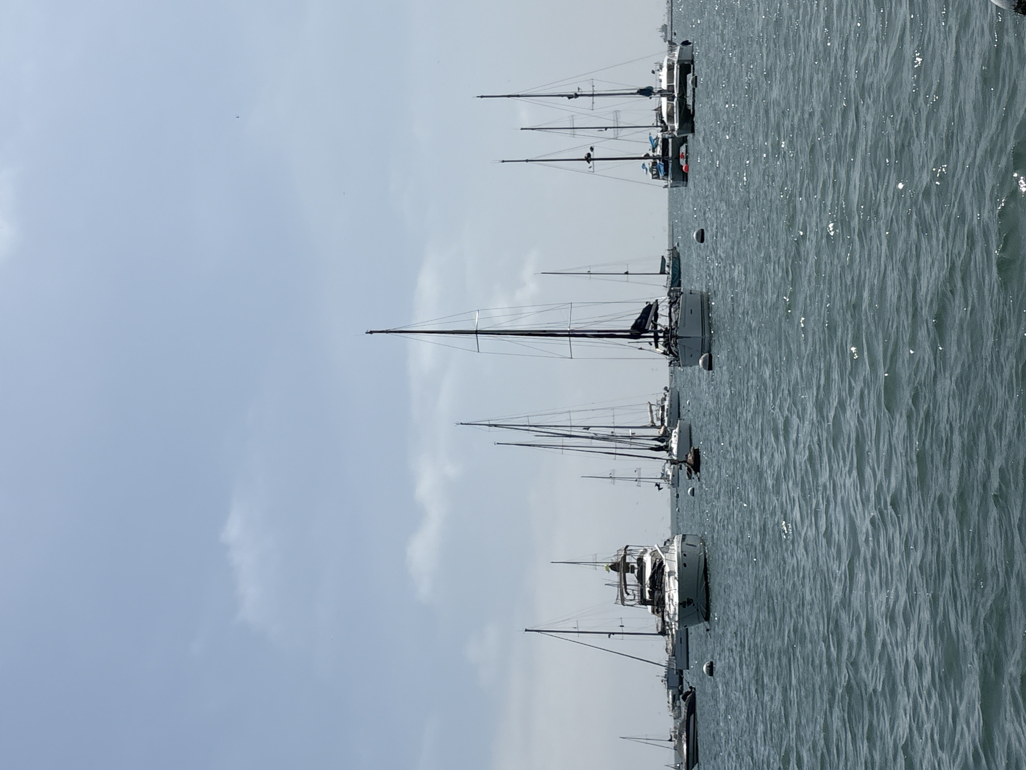 The image depicts a calm ocean scene with numerous sailboats anchored in the water. The boats have tall masts, and some have visible sails. The water surface reflects sunlight, creating a shimmering effect. The sky is overcast with light gray clouds. The perspective suggests the photo was taken from the shore, possibly in La Punta, Peru, based on the maritime environment characteristic of coastal towns. There are no people visible in the frame.