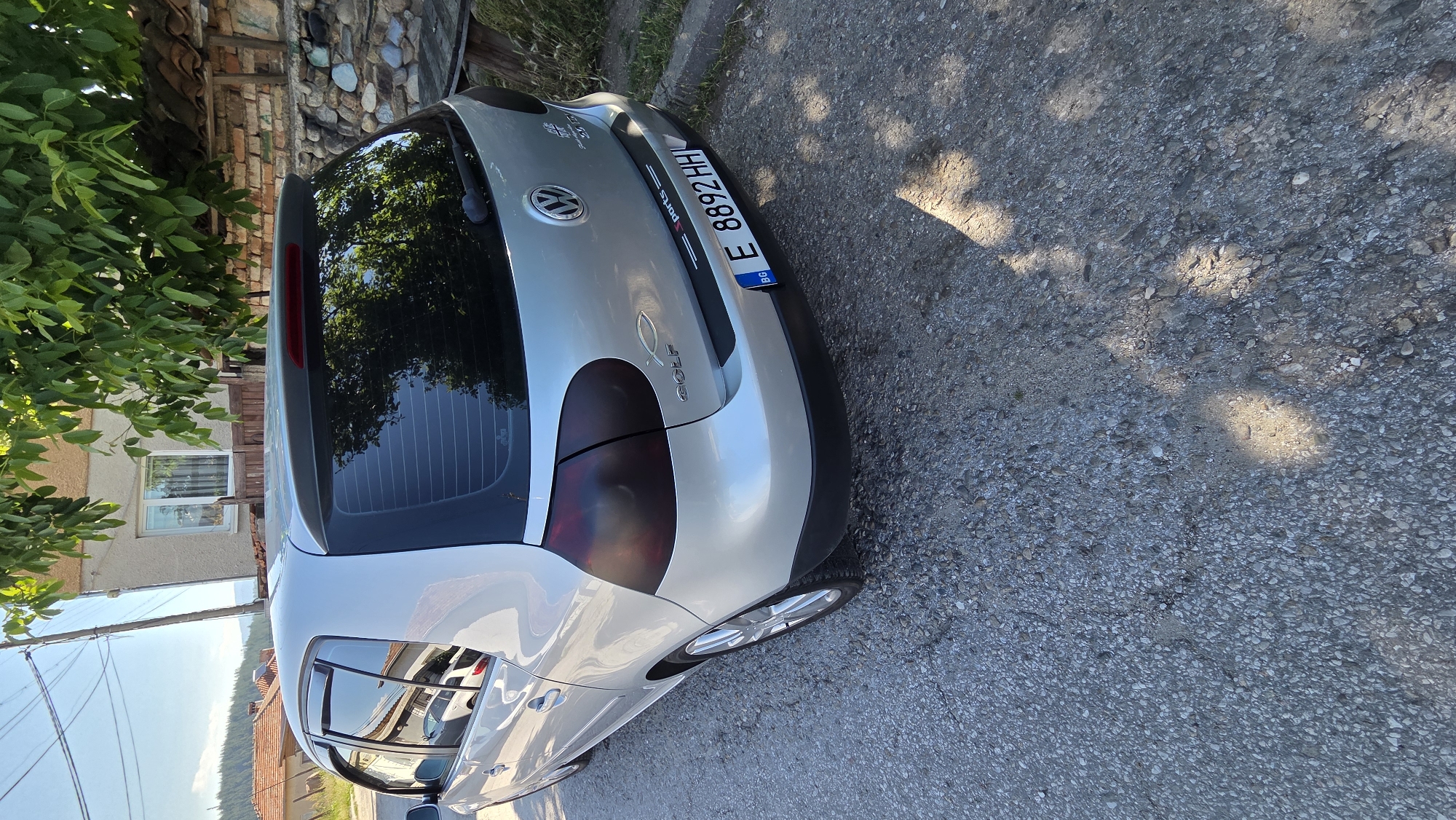 An outdoor, eye-level, medium shot captures the rear passenger side of a silver Volkswagen Golf parked on a gravel road. The car, positioned at an angle with its rear facing the right side of the frame, is the primary subject. Its tinted taillights and rear window are visible, reflecting the surrounding trees. The license plate, "E 8892 HH", with "BG" in a blue box on the left, is clearly displayed. A black strip above the license plate reads "ports" with a line on either side. The car's emblem, a circular Volkswagen logo, is centered on the trunk, and the word "GOLF" is visible below it, along with a small fish symbol.
The environment consists of a gravel road with scattered pebbles and some cracks. To the right and behind the car, a stone wall is present, topped with terracotta roof tiles. A rustic wooden table is partially visible next to the wall. Further in the background, a two-story building with a light-colored facade and a window is seen, partially obscured by lush green tree branches and leaves. The sky above is clear and blue, indicating daylight. Shadows from the trees are cast across the road and the car. No people are present in the image.