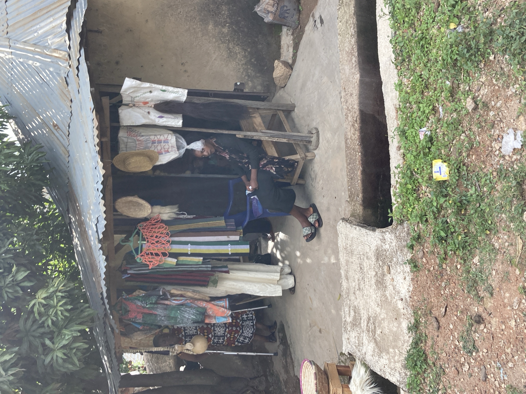 A woman is resting at her roadside stall in Jos, Nigeria. The stall is constructed from corrugated metal and wood, displaying various textiles and woven items. The woman, dressed in dark clothing with a patterned scarf, is seated in a blue plastic chair, her eyes closed. She appears to be napping or resting.
The stall is filled with colorful fabrics, including what look like traditional Nigerian attire. There are also hats and woven baskets visible. In front of the stall, there is a concrete drainage channel and a patch of overgrown greenery with some scattered debris. The scene suggests a warm, possibly sunny day, given the shadows and the typical weather in Nigeria. The overall impression is one of a quiet moment at a marketplace, where a vendor takes a break from selling her wares. The surroundings offer glimpses of a rural or semi-urban environment in Nigeria, with the natural vegetation and simple construction adding to the local context. No visible text is present in the image.