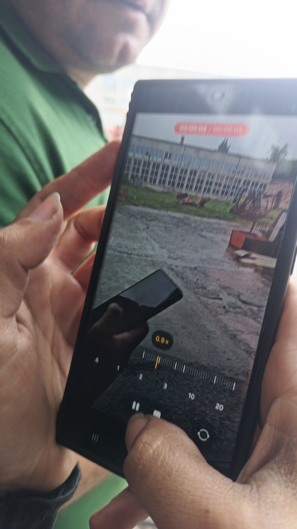 A person holds a smartphone displaying a paused video, with a timestamp of "00:00:04 - 00:00:09" visible on the screen. The video shows a building with multiple windows and what appears to be a yard or outdoor area, including what may be yard equipment, perhaps located in Karnobat, Bulgaria. The phone's screen interface shows a progress bar with markings for seconds, including "2," "3," "10," and "20," along with a pause button and a "0.9x" label, likely indicating the playback speed. Another phone is partially visible in the phone display. The person holding the phone is touching the screen, probably to control the video playback. Part of another person's green shirt is visible on the side.