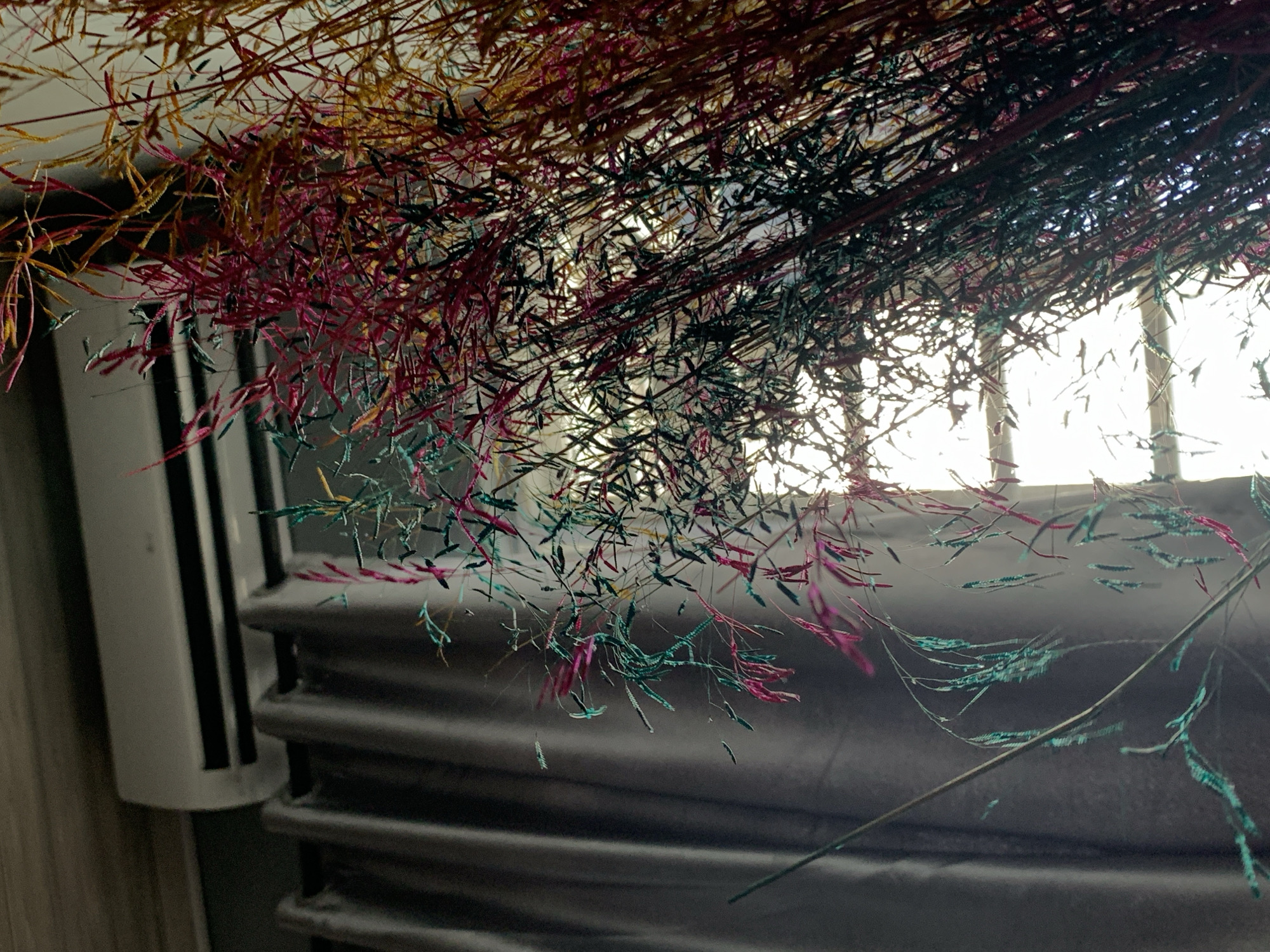 The image shows colorful decorative branches hanging in front of a window and a radiator. The branches are a mix of yellow, red, black, and blue-green colors. The background is a brightly lit window and a white radiator with black stripes. The radiator is in front of a grey wall. The location is likely a room in Ibasa, Nigeria. There are no people visible in the image.