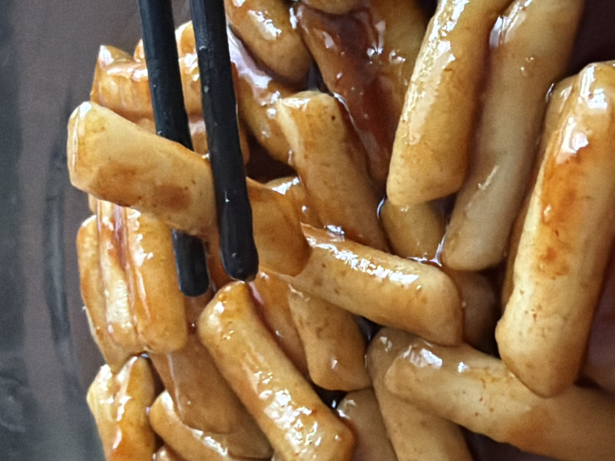 This close-up image presents a serving of food, most likely tteokbokki (Korean rice cakes), being handled by a pair of black chopsticks. The dish features numerous elongated, cylindrical pieces of golden-brown food, thoroughly coated in a thick, glossy, reddish-brown sauce. The sauce exhibits a viscous, glistening texture with prominent reflections, suggesting a rich and possibly sweet and spicy flavor.
In the upper left quadrant of the frame, one piece of the rice cake is held firmly by the simple black chopsticks, positioned as if being lifted to eat. The rice cakes have a smooth, slightly irregular surface, with their color varying from a deeper golden-brown to lighter, almost translucent shades where the sauce is thinner.
The background is dark and significantly blurred, creating a shallow depth of field that keeps the focus entirely on the food. This blurry background appears to be the interior of a dark bowl or container holding the dish. There are no individuals visible, and the image solely concentrates on the culinary subject and the action of eating or serving. No text is discernible within the frame. The scene lacks specific indicators for time of day, weather, or emotional context, but the nature of the food suggests a culinary experience, potentially within a Korean restaurant or home setting in Saint Petersburg, Russia.