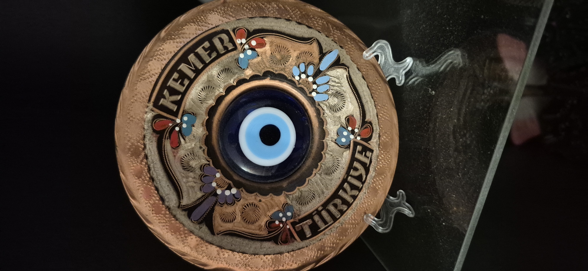The image shows a close-up of a decorative evil eye amulet plate, likely made of copper, featuring the words "KEMER" and "TÜRKIYE" in an arched inscription. The amulet itself is a circular blue glass eye with concentric white and black rings. Decorative floral and leaf motifs in red, blue, and purple are painted around the eye. The plate is supported by two transparent plastic stands on a dark, reflective surface, possibly a glass table. The background is black. The scene is a still life, and there is no indication of the city of Baden-Baden, Germany.