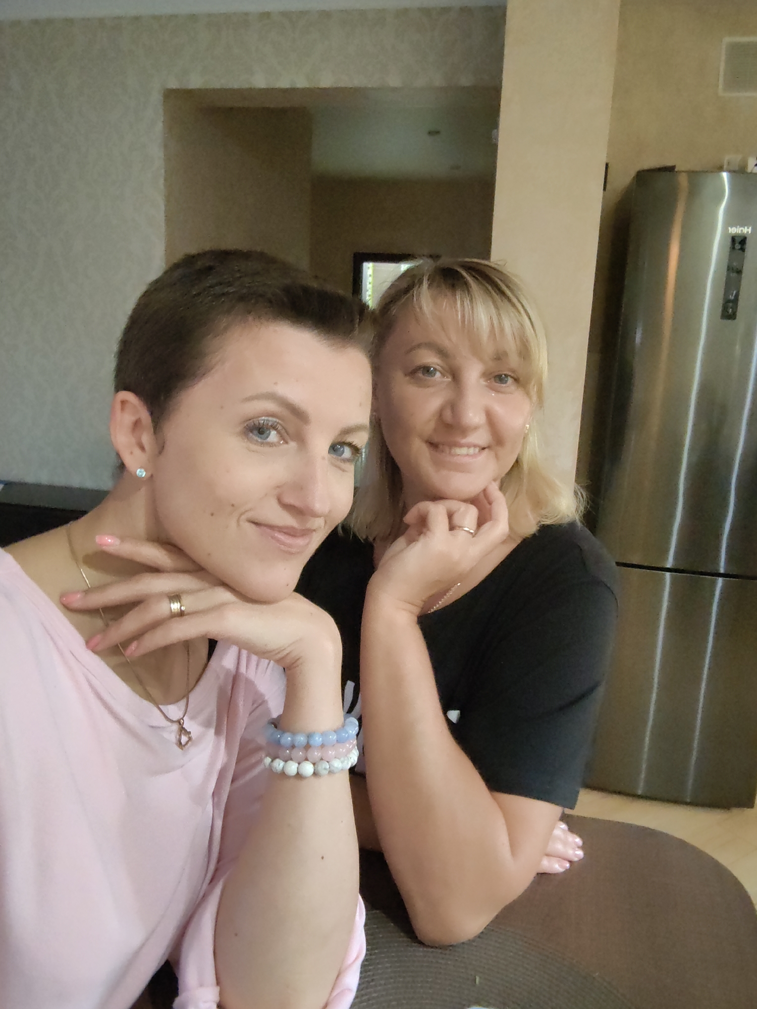The image depicts two women in what appears to be a home environment, likely a kitchen given the presence of a stainless steel refrigerator on the right side of the frame. The woman in the foreground, on the left, has short, dark hair and is wearing a pink top. She has her hand resting on her chin, adorned with rings and bracelets. Her gaze is directed towards the camera. The second woman, on the right, has blonde hair and is wearing a black top. She is also looking towards the camera, with her hand near her chin. The background shows a neutral-colored wall and a doorway. The overall lighting suggests an indoor setting.