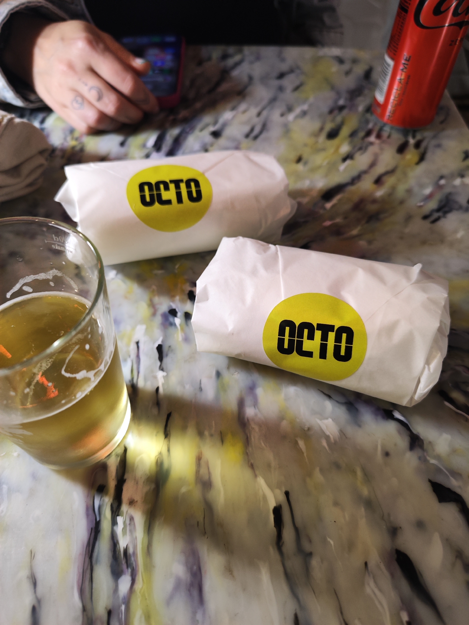 The image shows a table in Lisbon, Portugal. On the table are two wrapped sandwiches with yellow circular "OCTO" stickers, a glass of beer, and a can of Coca-Cola. A person's hand holding a phone is visible in the background. The table has a marbled, multi-colored surface.
