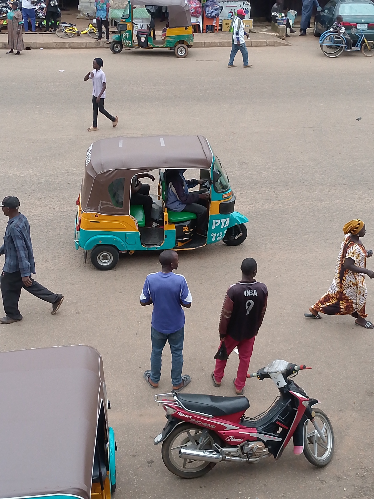 This is a street scene in Bukuru, Nigeria, on a paved road with a brown and teal auto-rickshaw in the foreground. A red and black motorcycle is parked to the right of the auto-rickshaw. To the left of the auto-rickshaw, a man in a checkered blue and gray shirt and dark pants walks away from the camera. Two men stand in the middle ground, one in a blue shirt and jeans, the other in a black shirt with "OBA 9" on the back and red pants. Further back, a woman in a patterned dress walks to the right. A young man in a white shirt and black pants walks across the road from left to right, further in the background. In the far background, a green car and a three-wheeled vehicle with a green canopy are visible, along with other pedestrians and market stalls. The overall impression is of a busy, everyday street scene.