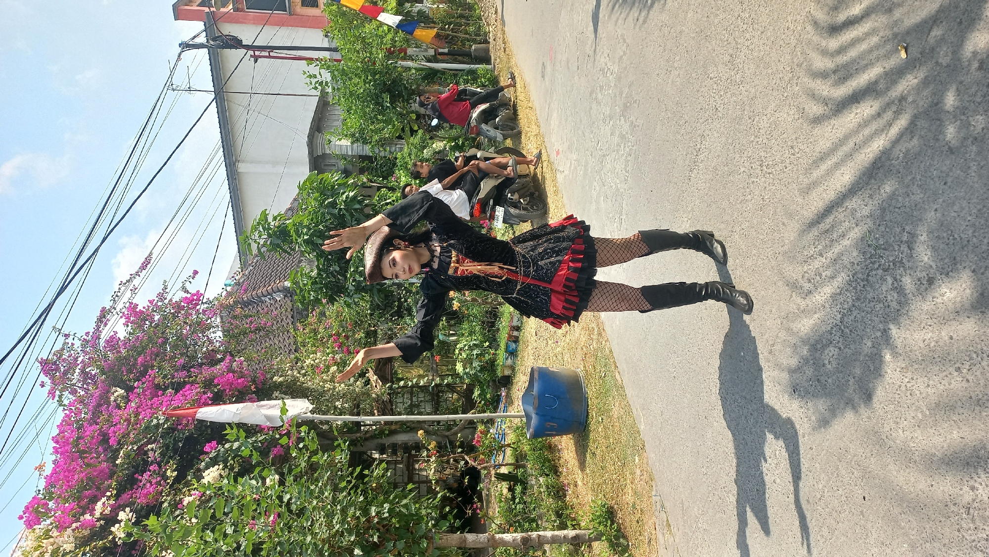In Kedungmegarih, Indonesia, a young woman in a black and red pirate-themed costume with a feathered hat and fishnet stockings stands in the middle of a paved road, her arms raised. She is wearing black knee-high boots. Behind her, several people are visible. Two men are seated on a motorcycle, and another person, possibly a woman, is on a separate motorcycle further down the road. The background features lush green trees, a house with a tiled roof, and a red and white Indonesian flag on a pole amidst pink and white bougainvillea flowers. The scene is illuminated by bright sunlight, casting shadows on the road.