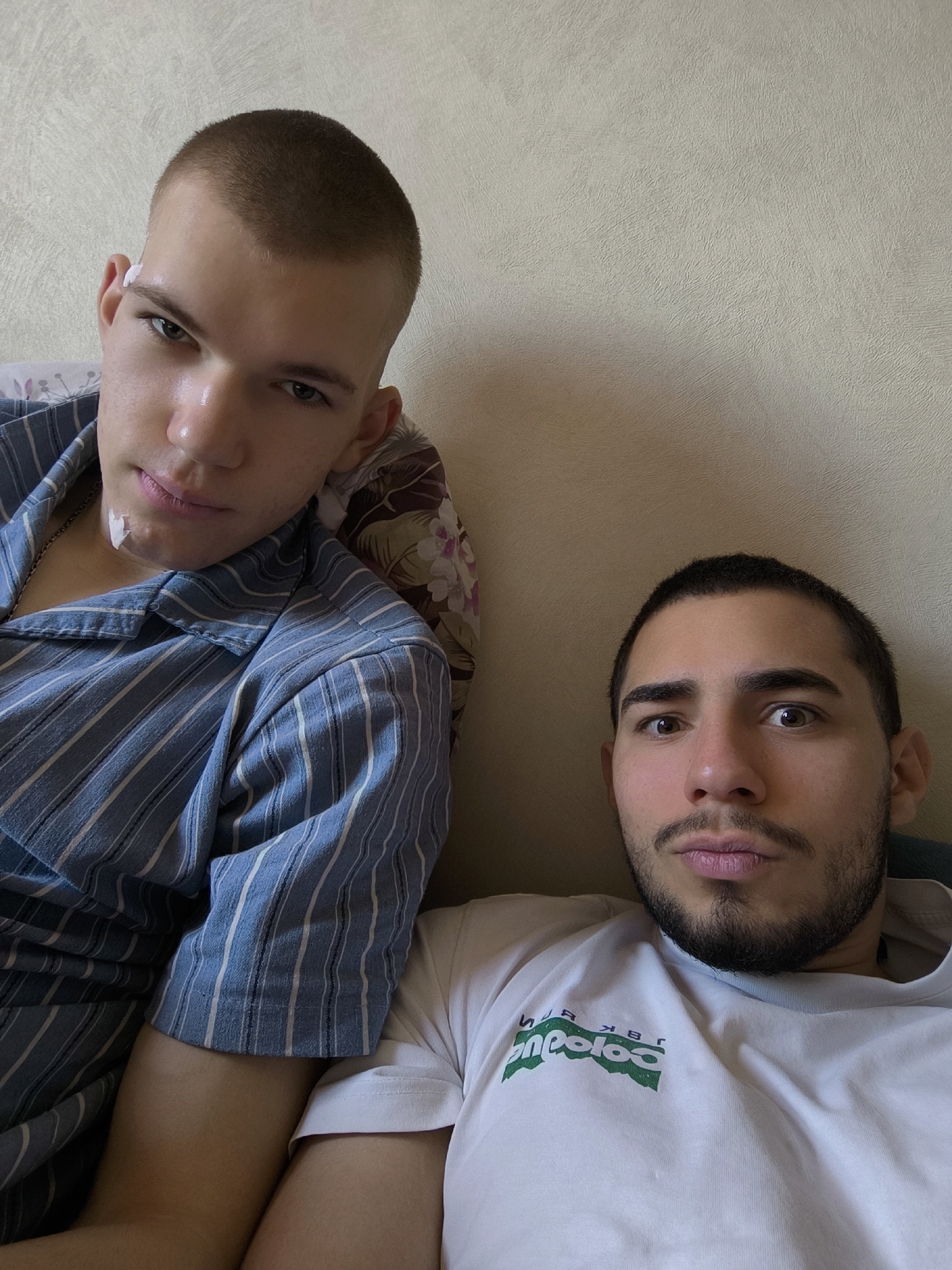 An eye-level indoor portrait captures two young men posing for a selfie, likely within a home environment in Schiltigheim, France. Both subjects gaze directly at the camera with neutral to slightly serious expressions.
The man on the left has a light complexion, very short light brown hair, and light-colored eyes. He is wearing a blue and white striped long-sleeved button-up shirt and is propped against a pillow with a floral design. A small white piece of material, possibly medical tape or cotton, is visible behind his left ear and another on his chin/neck area.
The man on the right features a darker complexion, short dark hair, dark eyes, and a trimmed beard and mustache. He wears a white short-sleeved T-shirt with a green, mirrored logo on the chest. The visible text, when reversed, appears to include "BRK" and "Coloco" or "Coloc".
The background is a simple, light-colored wall with a subtle textured finish. The lighting is soft and even, consistent with natural daylight indoors. The overall scene suggests a relaxed and casual setting.