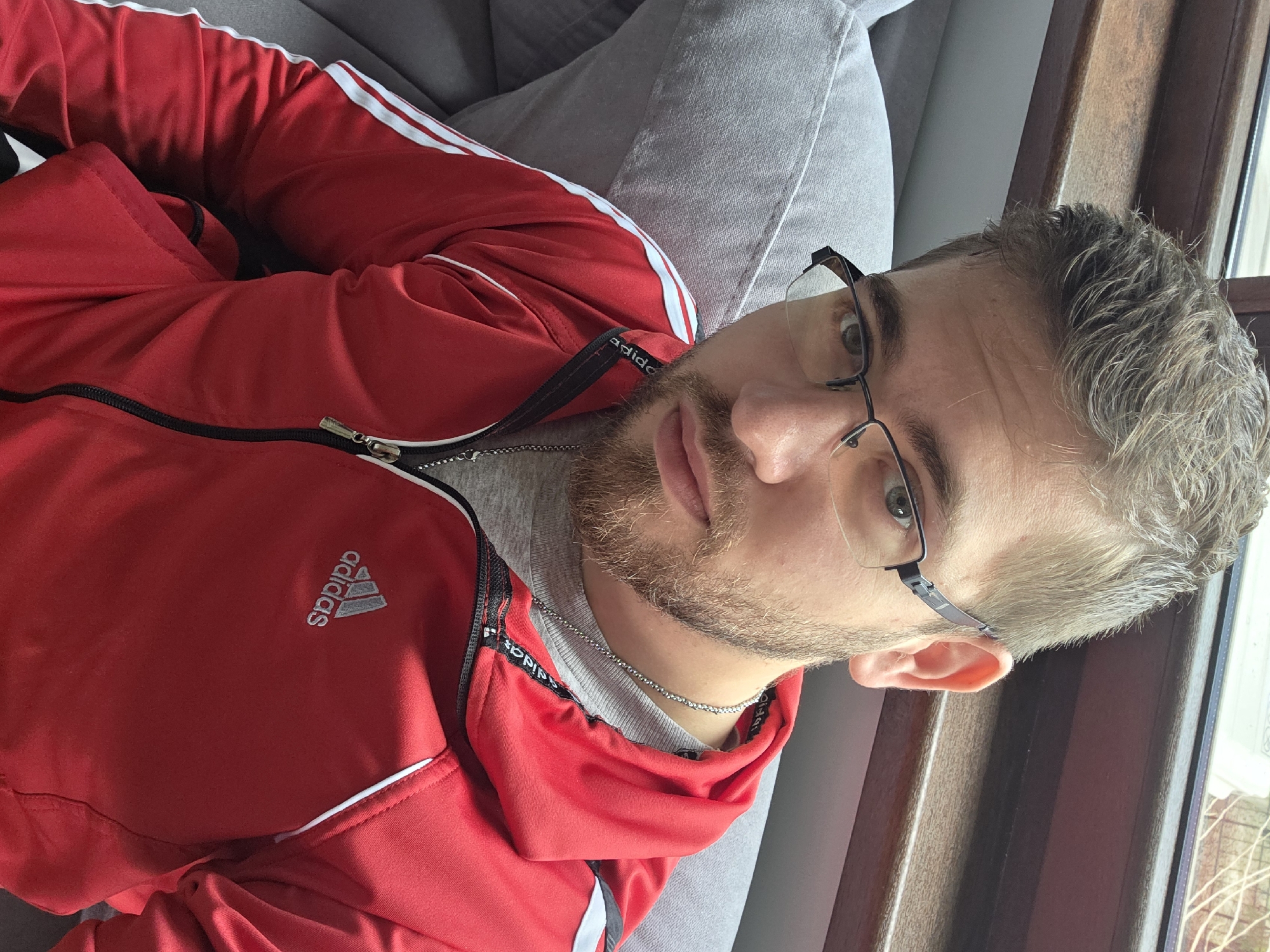 A fair-skinned male with short, light brown hair and a trimmed beard is reclined on a light gray sofa. He wears glasses with dark frames and a red Adidas tracksuit with white stripes. The location is indeterminable based on the image; there is no evidence to suggest the image was taken in Yagodovo, Bulgaria. A window with a brown frame is partially visible behind him. He is wearing a silver chain.