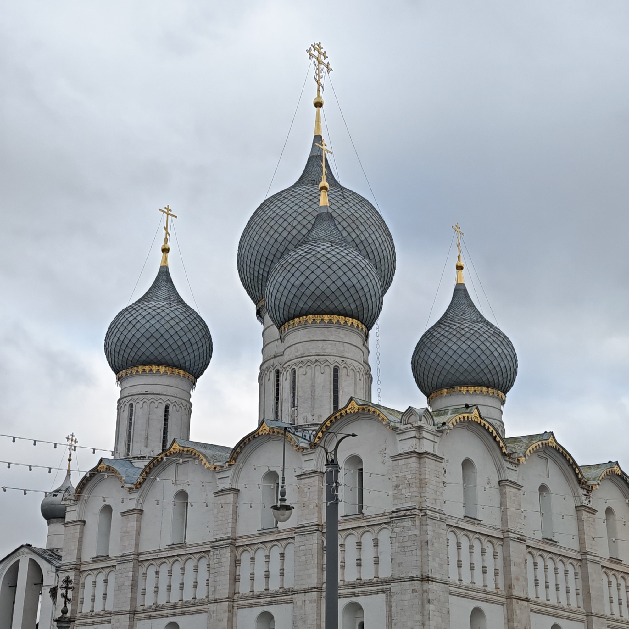 The image shows the Cathedral of the Dormition, located in Rostov, Russia. It features five dark, onion-shaped domes, each topped with a golden cross. The cathedral is constructed of white stone and exhibits ornate architectural details, including arches and decorative gold trim along the rooflines. A street lamp is visible in the foreground, and string lights are strung in the background. The sky is overcast, indicating a cloudy day.